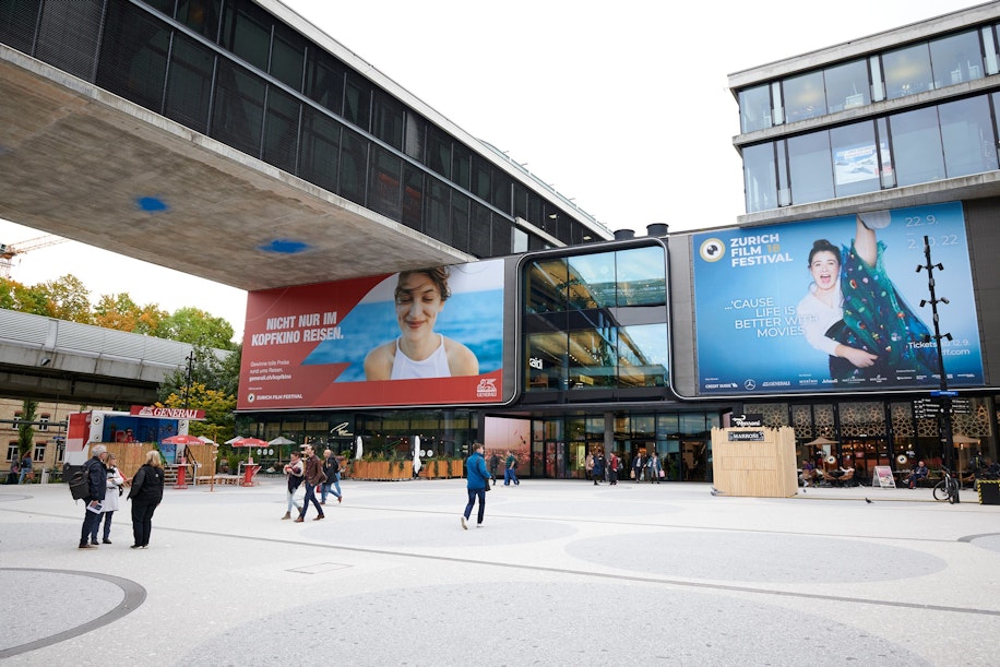 Ein moderner Stadtplatz mit flanierenden Menschen und großen Werbetafeln an zeitgenössischen Gebäuden, darunter ein Plakat des Zürcher Filmfestivals und ein weiteres mit einer lächelnden Frau.