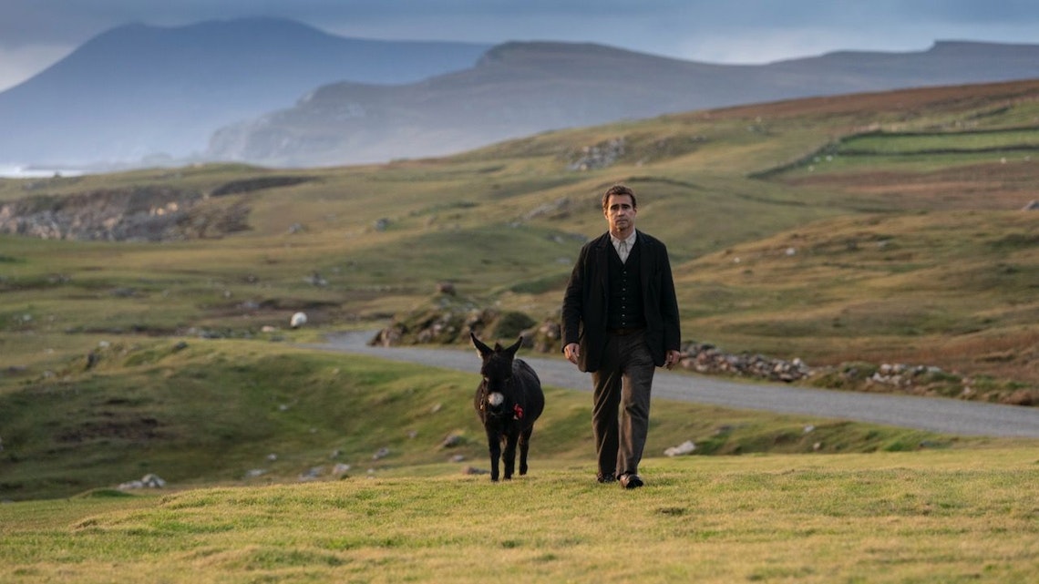 A man in dark clothing walks beside a small donkey on a grassy, hilly landscape with a winding road and mountains in the distance under a cloudy sky.