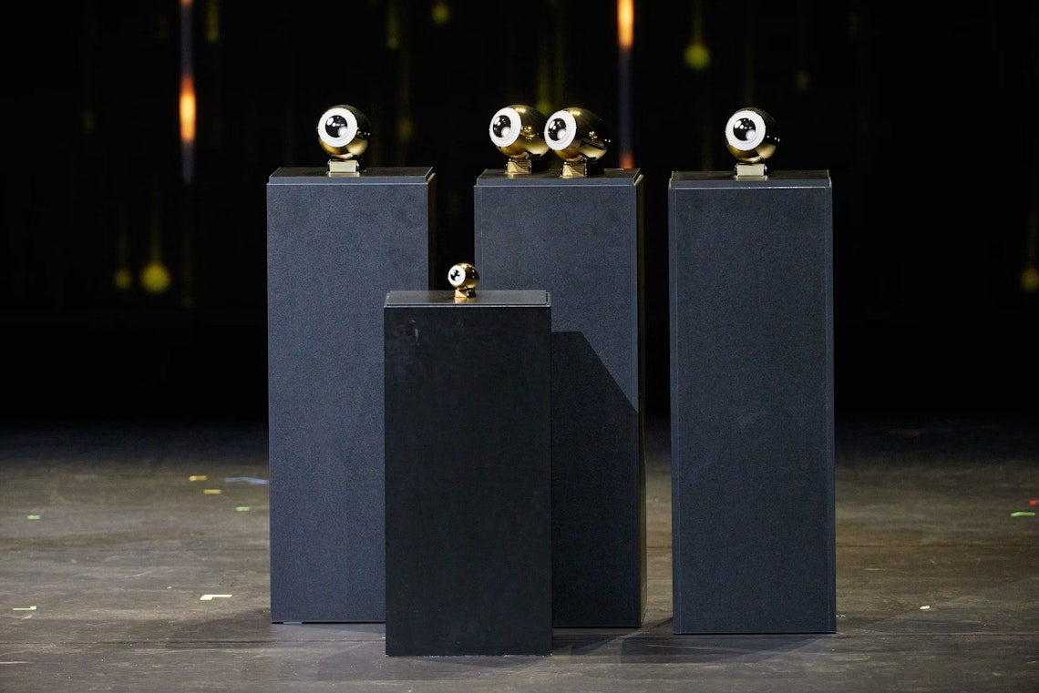 Four black podiums of varying heights display four gold and black trophies shaped like eyes, with a dark, blurred background and soft lights behind them.