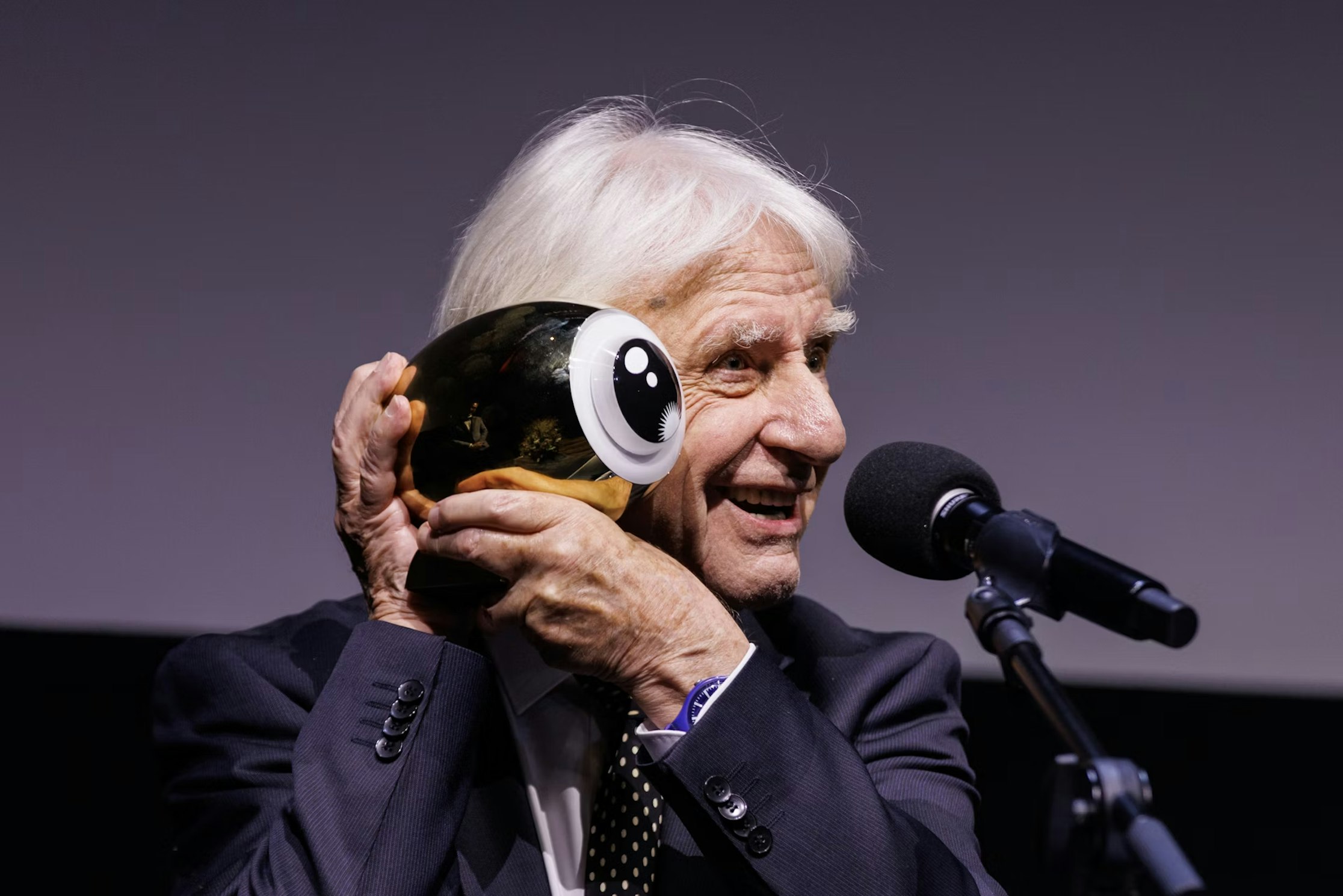 Ein älterer Mann im Anzug lächelt und hält eine glänzende, augenförmige Trophäe an sein Ohr, während er auf der Bühne in ein Mikrofon spricht.
