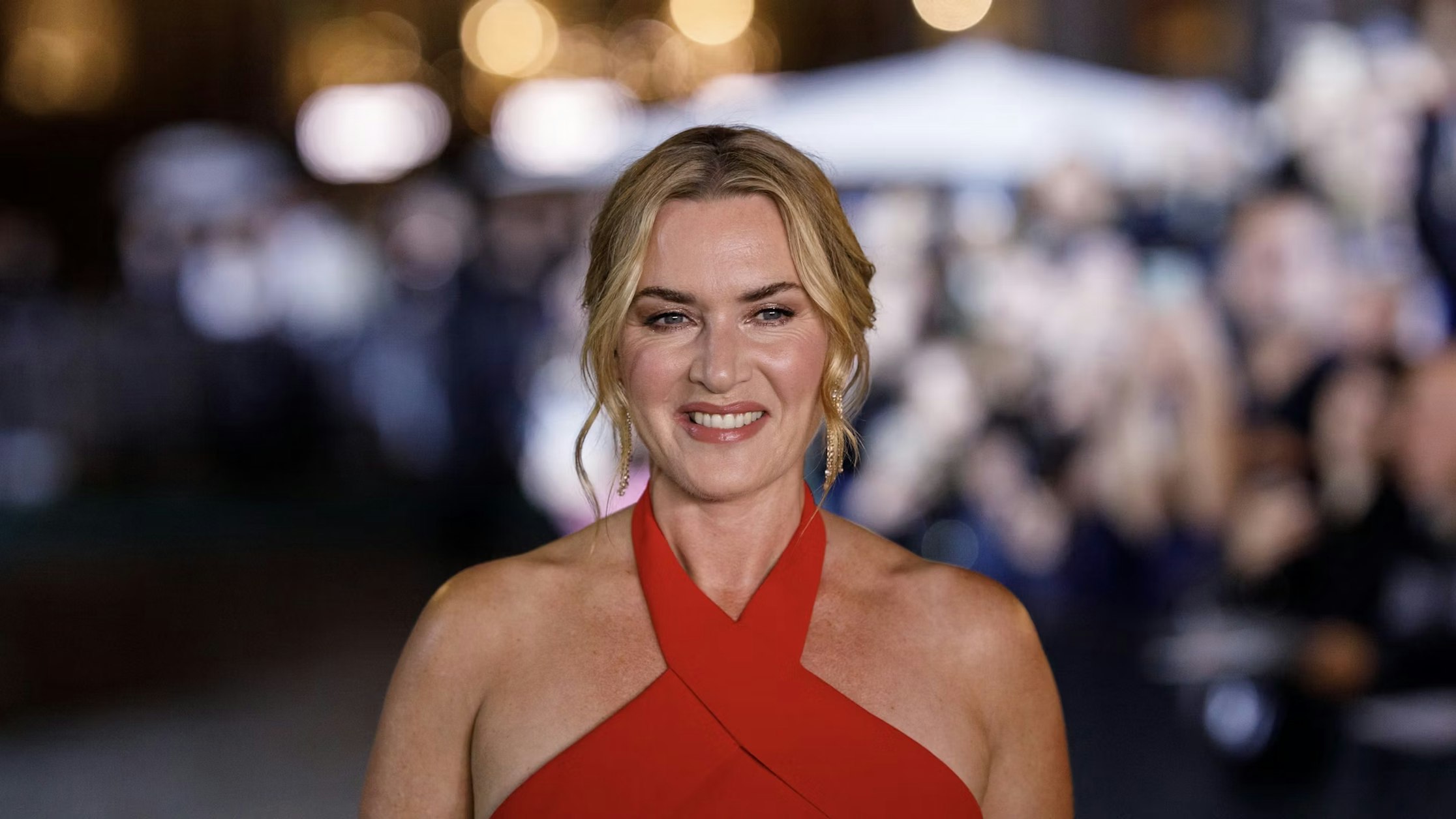 Eine Frau mit welligem blondem Haar und einem roten Neckholder-Kleid lächelt bei einer Veranstaltung mit verschwommenem Licht und einer Menschenmenge im Hintergrund.