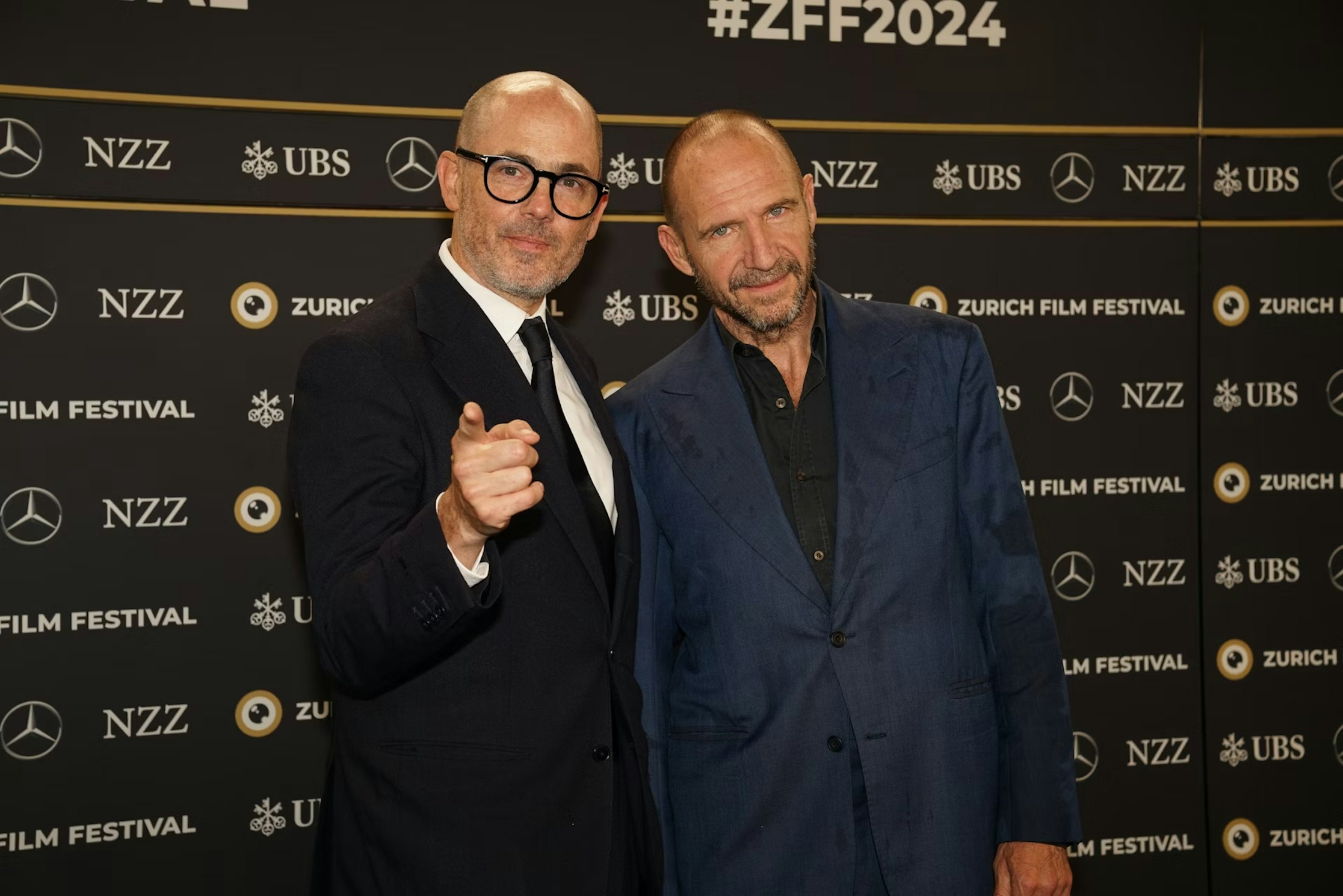 Zwei Männer in Anzügen posieren gemeinsam am Zurich Film Festival vor einer schwarzen Kulisse mit Sponsorenlogos und #ZFF2024. Ein Mann zeigt in die Kamera, beide lächeln.