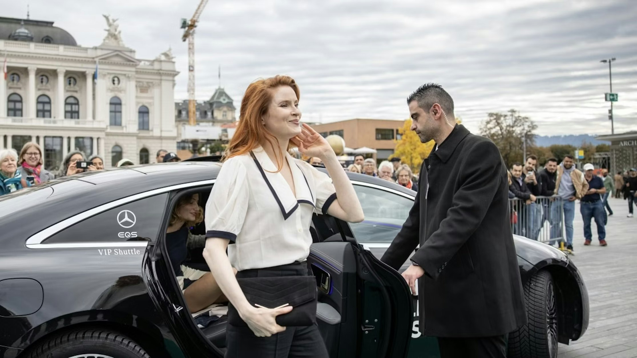 Eine rothaarige Frau steigt aus einem schwarzen Mercedes-Benz. Ein Mann hält ihr die Tür auf. Hinter einer Absperrung stehen Schaulustige. Im Hintergrund sind historische Gebäude und ein Baukran zu sehen.