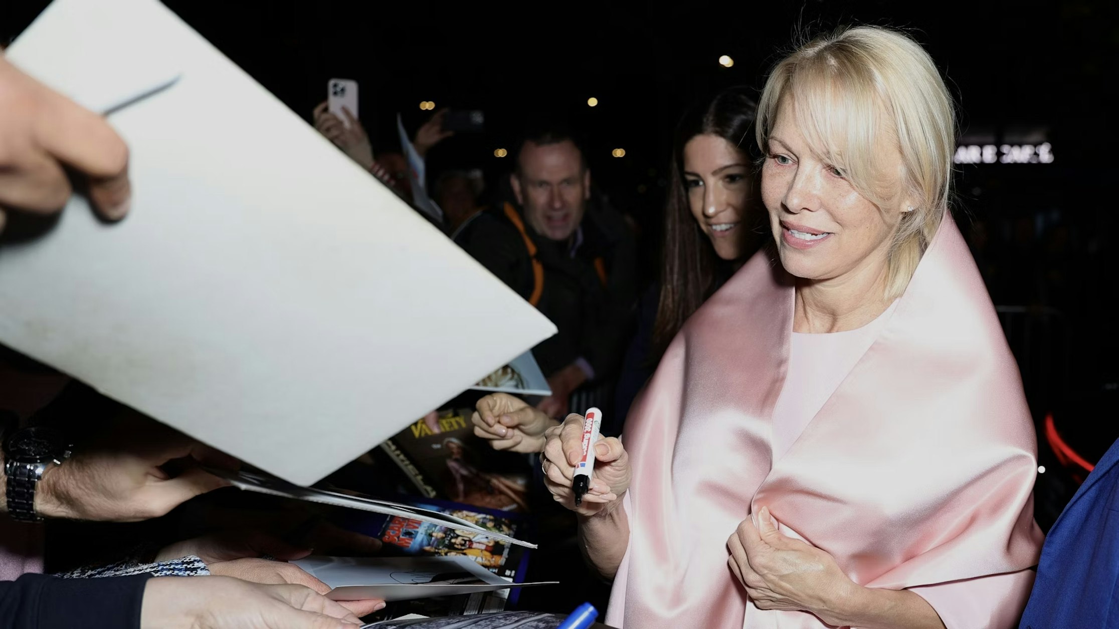 Eine Frau mit einem hellrosa Schal lächelt, während sie abends Autogramme für Fans gibt. Um sie herum halten Menschen Fotos und Stifte hoch, im Vordergrund sind mehrere Hände zu sehen.