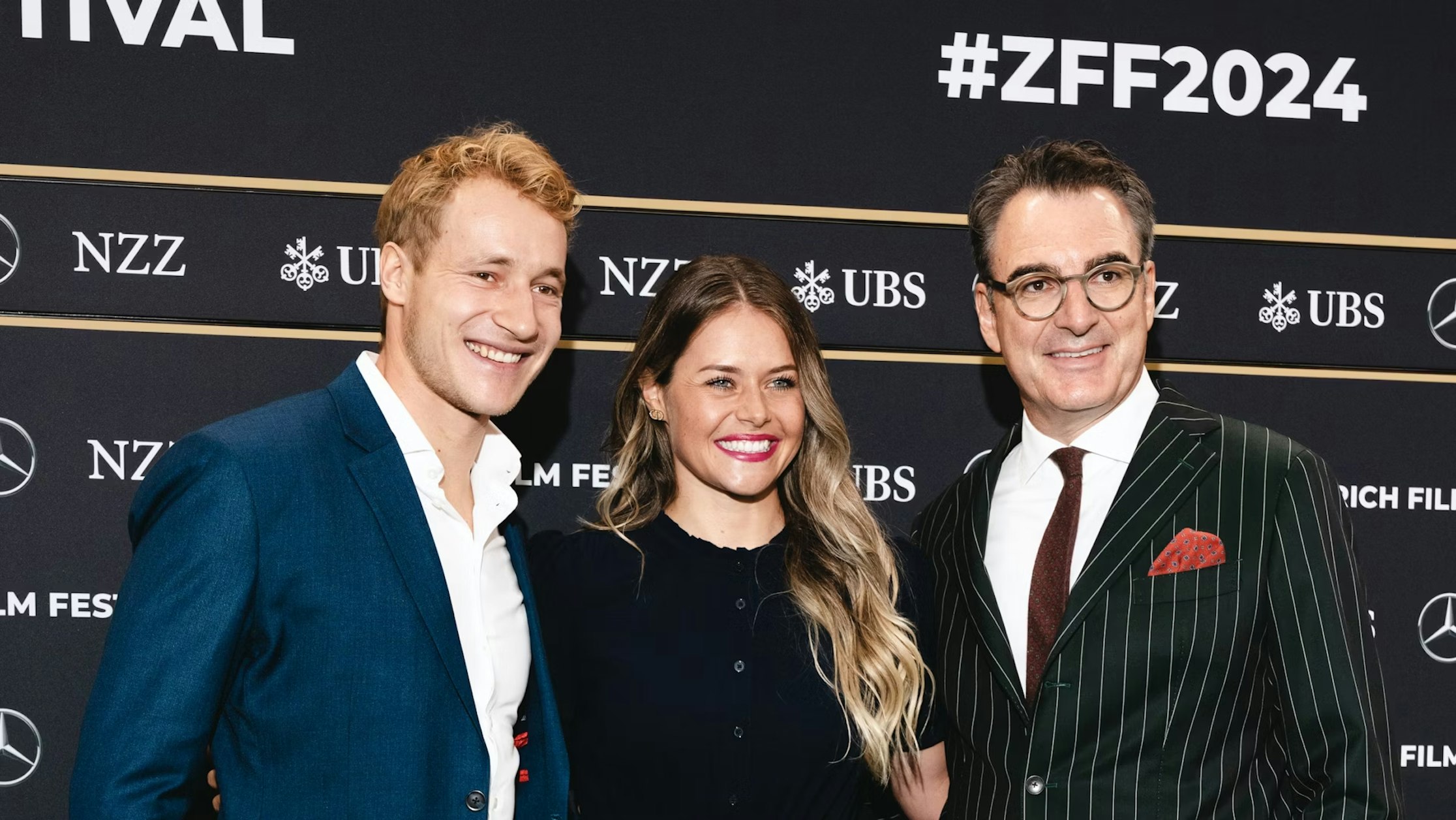 Drei Personen stehen lächelnd vor der Kulisse des Zurich Film Festival 2024. Der Mann links trägt einen blauen Anzug, die Frau in der Mitte trägt Schwarz und der Mann rechts trägt einen Nadelstreifenanzug und eine Brille.