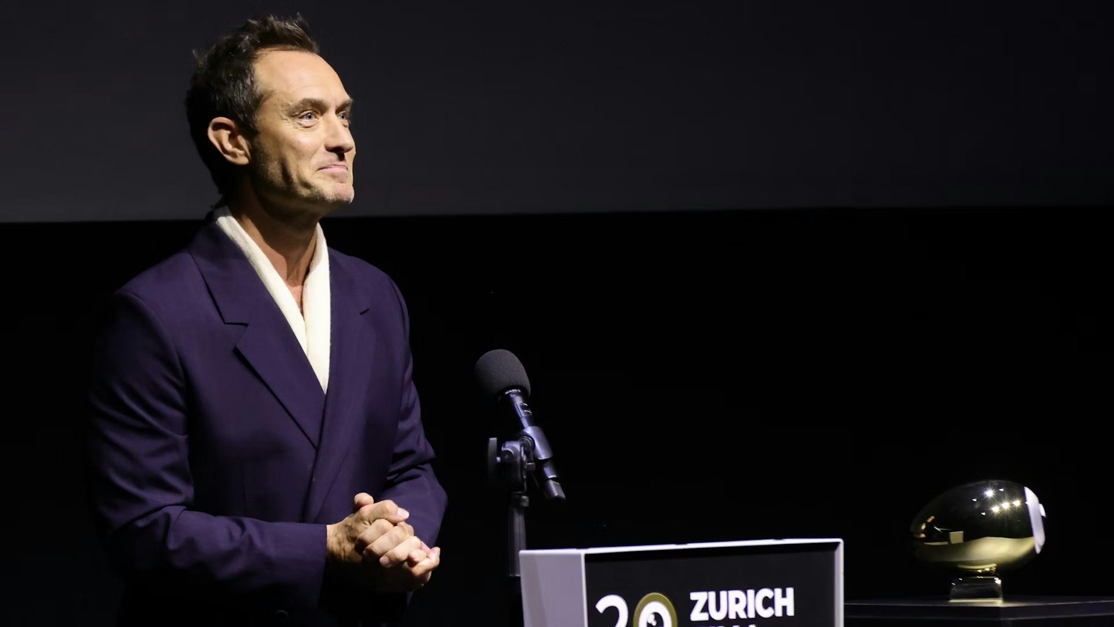 Ein Mann im dunklen Anzug steht an einem Podium mit der Aufschrift „Zürich“ und spricht in ein Mikrofon. Neben ihm liegt auf dem Tisch eine goldene Auszeichnung, dahinter ein dunkler Hintergrund.