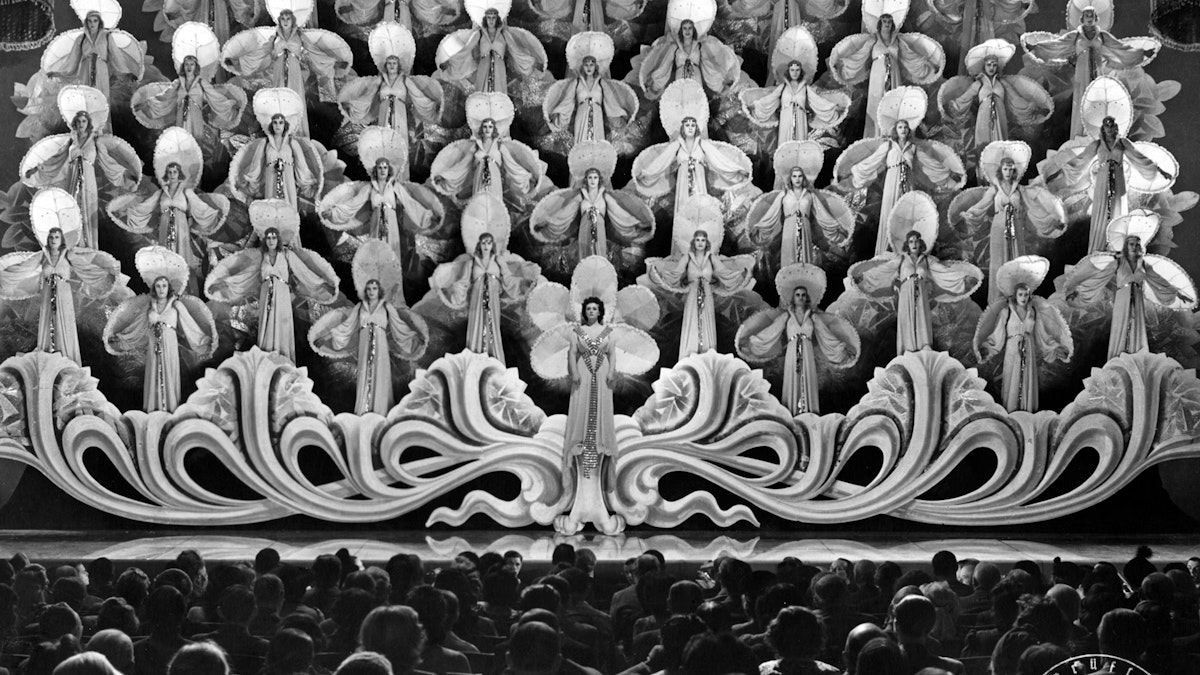 A large group of women in elaborate costumes with fans stand on tiered platforms on a stage, facing an audience. Ornate decorations and dramatic lighting create a grand, theatrical scene.