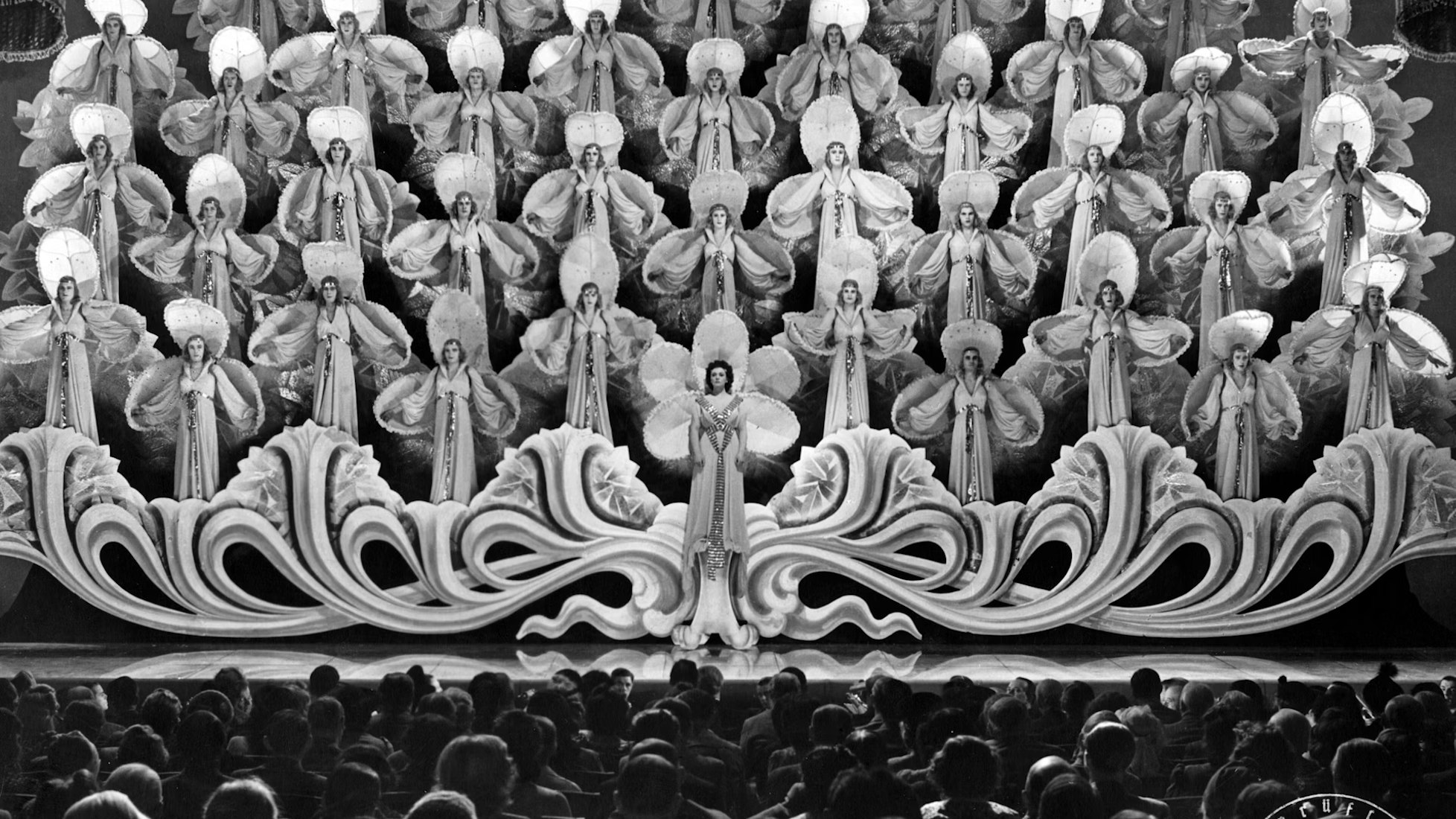 A large group of women in elaborate costumes with fans stand on tiered platforms on a stage, facing an audience. Ornate decorations and dramatic lighting create a grand, theatrical scene.