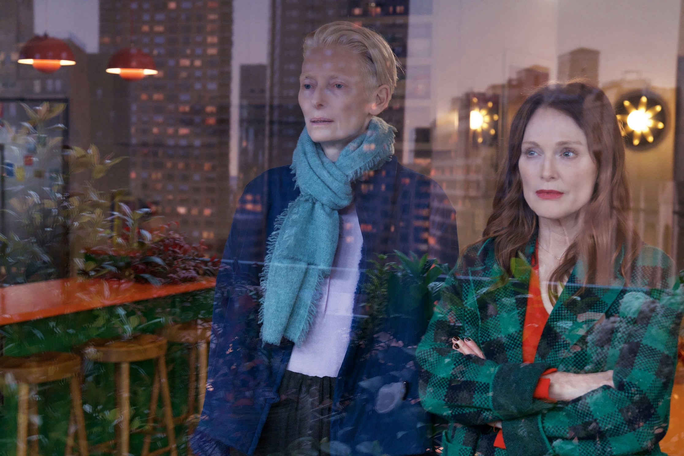 Zwei Frauen stehen drinnen an einem Fenster. Ihre Spiegelbilder verschmelzen mit der urbanen Stadtlandschaft und einem gemütlichen Barbereich mit Pflanzen und Barhockern. Die eine trägt einen blauen Schal, die andere einen grünkarierten Mantel. Beide blicken nachdenklich.