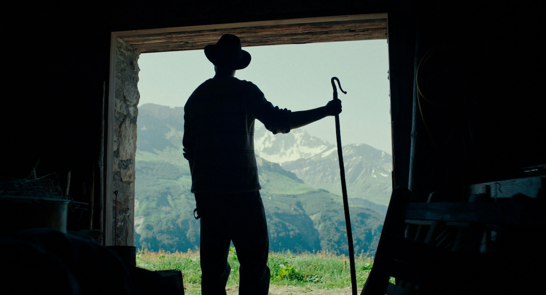 Die Silhouette einer Person mit Hut und Stab steht in einem Türrahmen und blickt auf sonnenbeschienene Berge und grasbewachsene Hügel in der Ferne.