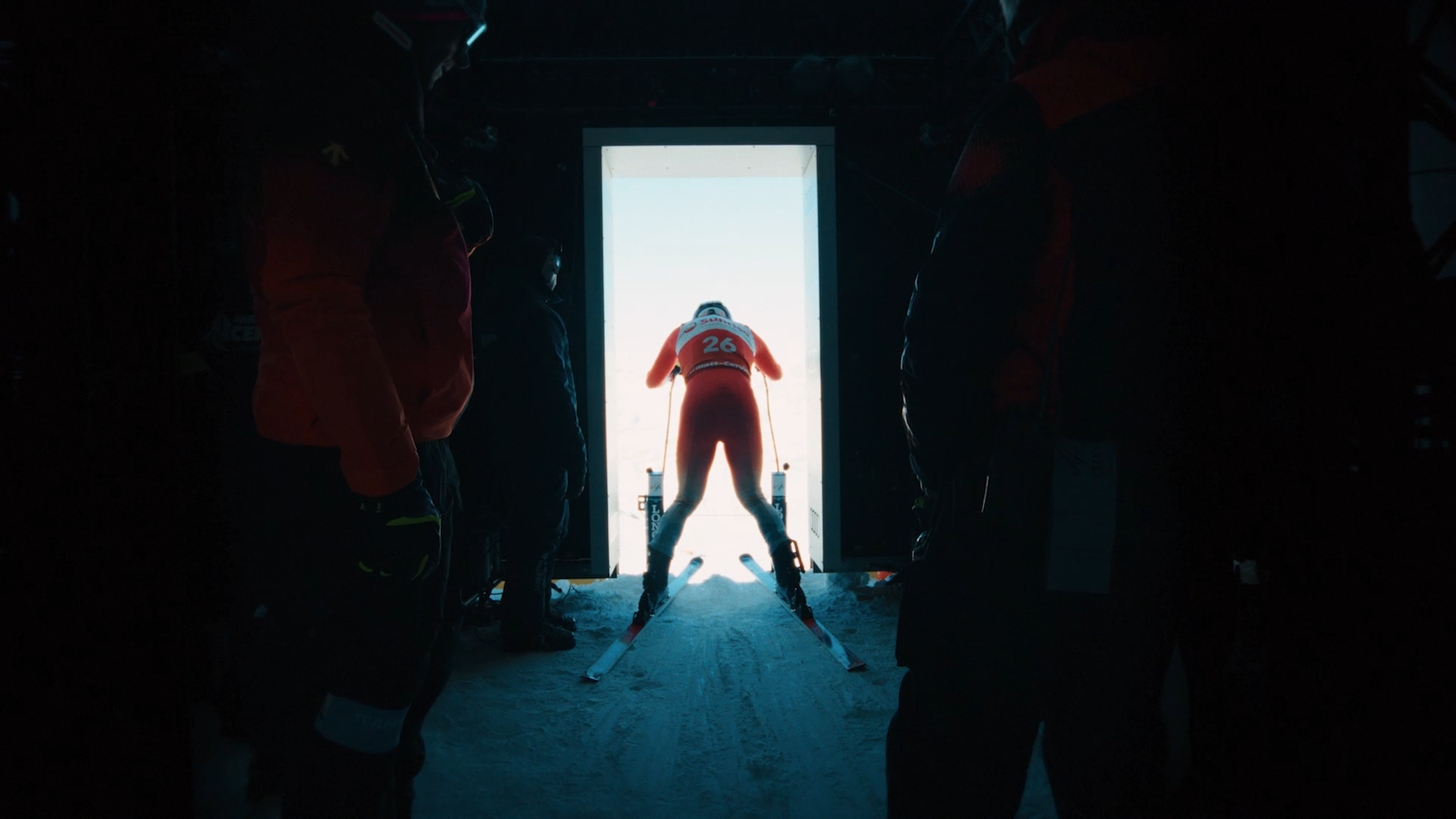 Ein Skifahrer in einem roten Anzug mit der Nummer 26 bereitet sich darauf vor, aus einem Hauseingang, flankiert von zwei schattenhaften Gestalten in Winterkleidung, in eine helle Schneelandschaft hinauszufahren.