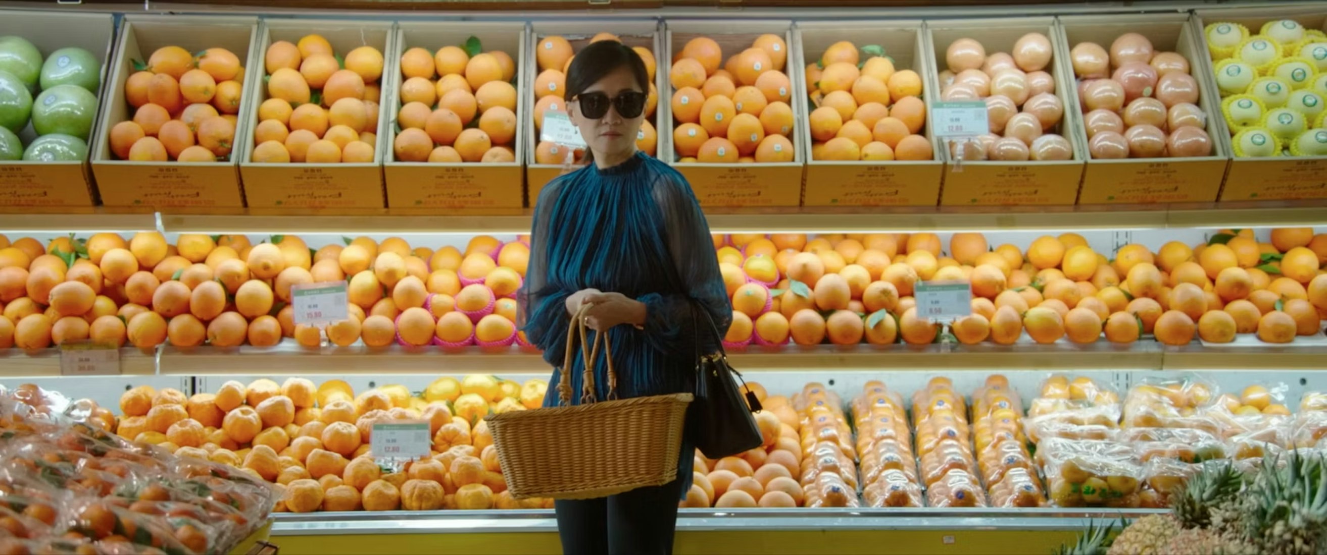 A woman wearing sunglasses and a blue scarf stands in a grocery store produce aisle, holding a wicker basket. Shelves behind her are filled with neatly arranged oranges and other citrus fruits.