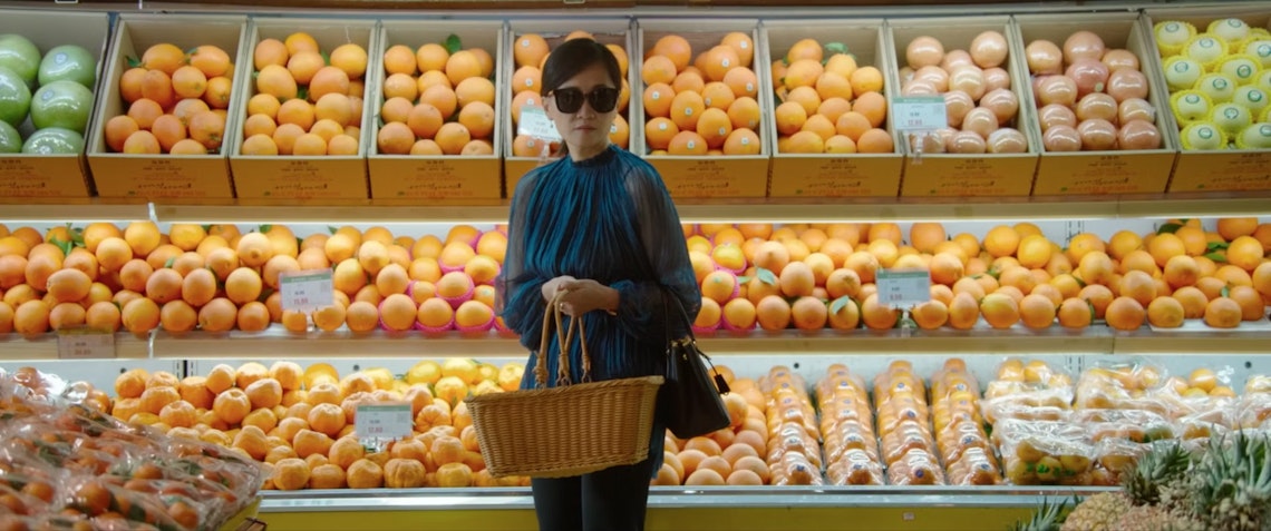 A woman wearing sunglasses and a blue scarf stands in a grocery store produce aisle, holding a wicker basket. Shelves behind her are filled with neatly arranged oranges and other citrus fruits.