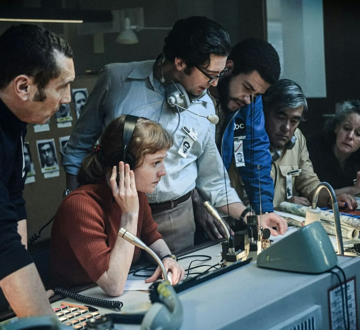 Six people gather around a desk with audio equipment and documents, appearing focused and engaged in discussion. Some wear headsets and ID badges, suggesting a newsroom or control room setting.