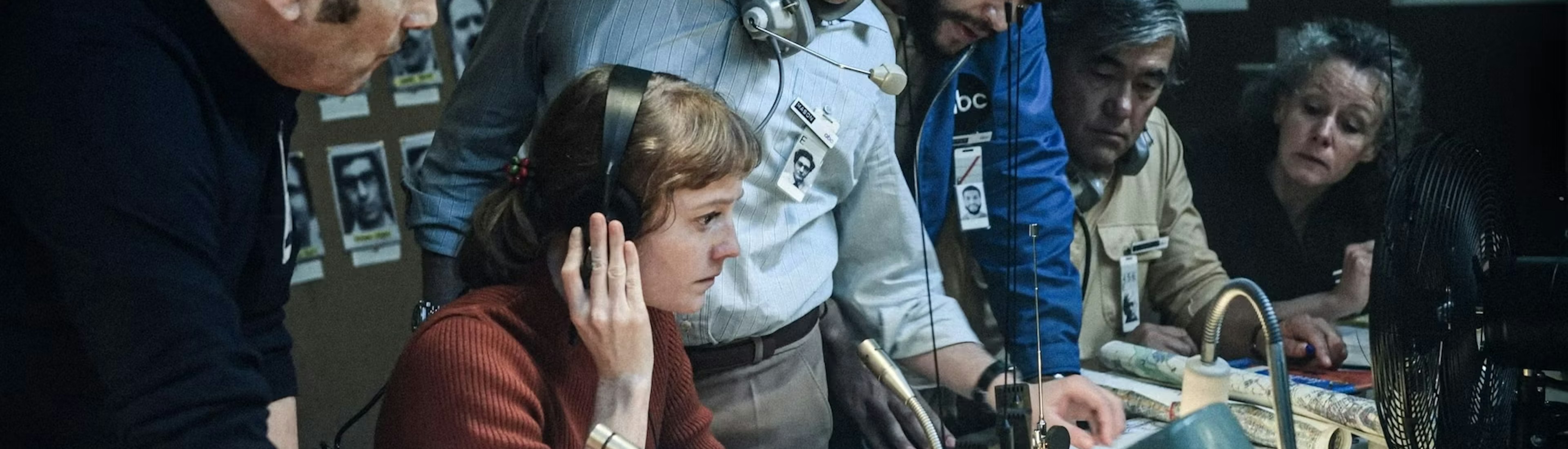 Six people gather around a desk with audio equipment and documents, appearing focused and engaged in discussion. Some wear headsets and ID badges, suggesting a newsroom or control room setting.