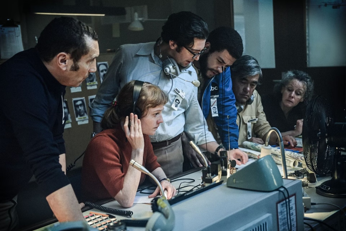 Six people gather around a desk with audio equipment and documents, appearing focused and engaged in discussion. Some wear headsets and ID badges, suggesting a newsroom or control room setting.