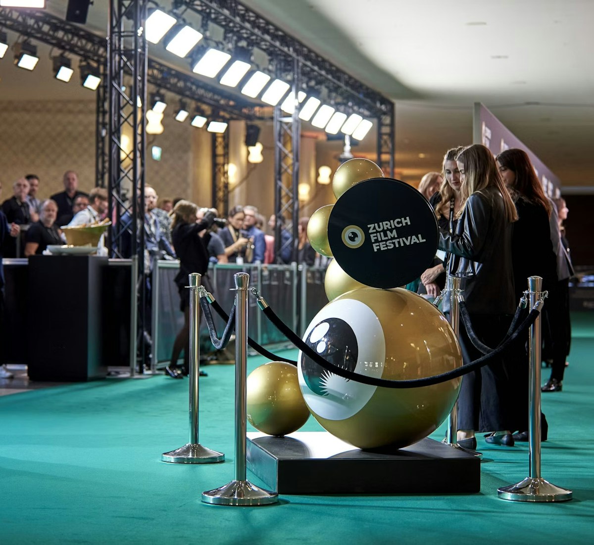 People stand on a green carpet at the Zurich Film Festival, with photographers behind barriers and a large gold festival logo sculpture in the foreground, under bright event lighting.