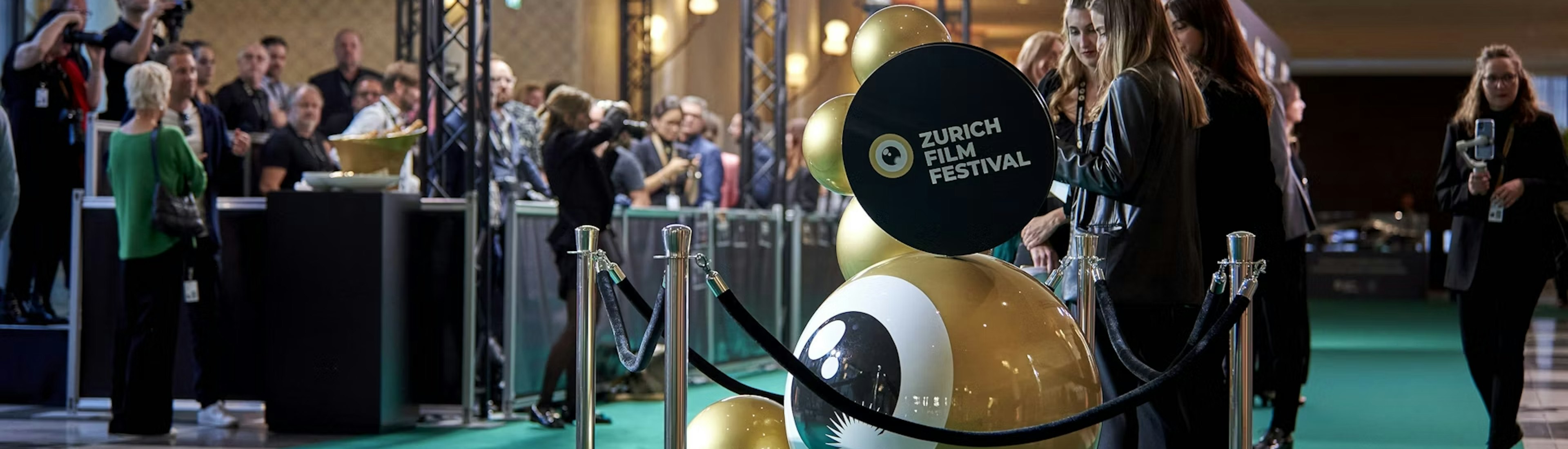 People stand on a green carpet at the Zurich Film Festival, with photographers behind barriers and a large gold festival logo sculpture in the foreground, under bright event lighting.