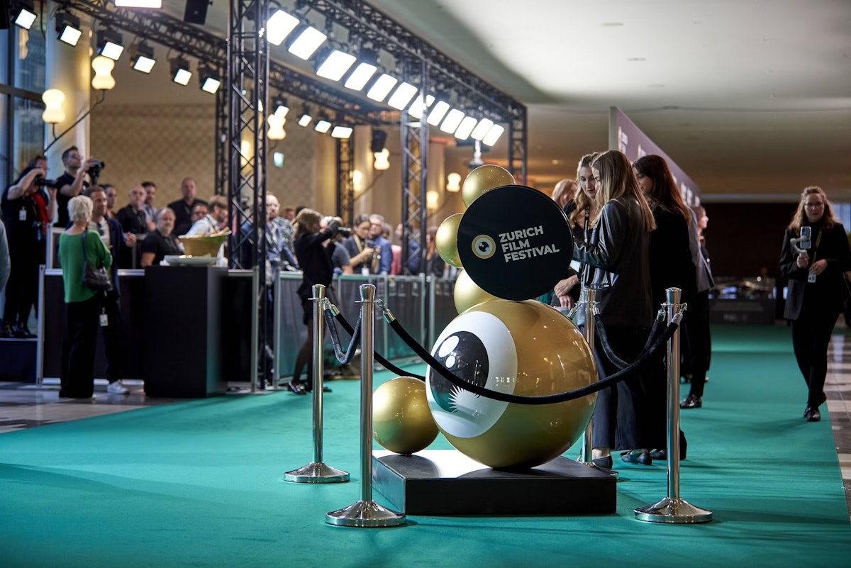People stand on a green carpet at the Zurich Film Festival, with photographers behind barriers and a large gold festival logo sculpture in the foreground, under bright event lighting.