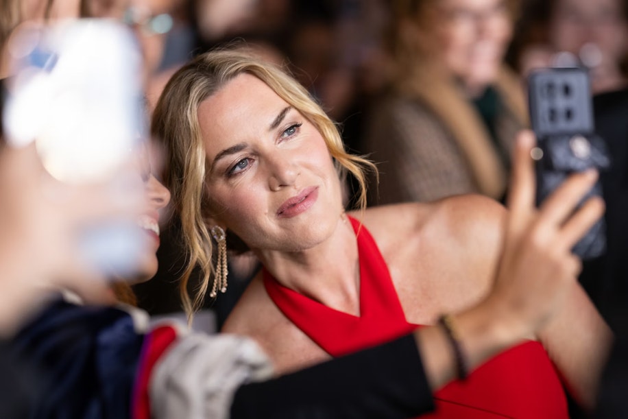 Eine Frau in einem roten Kleid lächelt, während sie bei einer gut besuchten Veranstaltung mit einem Fan für ein Selfie posiert. Sie hat blondes, welliges Haar und trägt baumelnde Ohrringe. Der Hintergrund ist unscharf, andere Personen sind zu sehen.