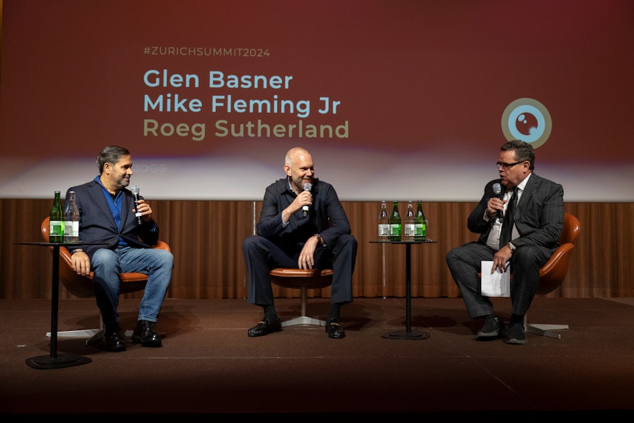 Drei Männer sitzen auf der Bühne vor einem Bildschirm, auf dem die Namen Glen Basner, Mike Fleming Jr. und Roeg Sutherland beim Zurich Summit 2024 zu sehen sind. Auf den Tischen daneben stehen zwei Handmikrofone und Wasserflaschen.