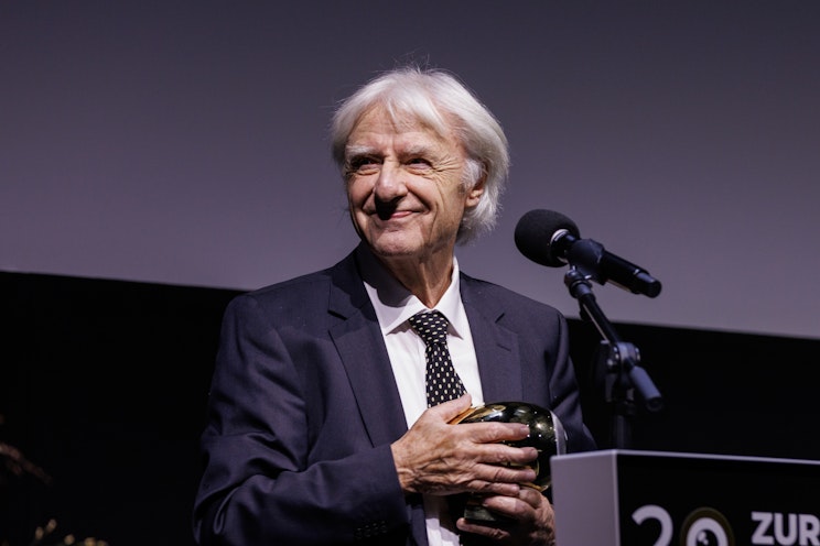 Ein älterer Mann in einem dunklen Anzug und einer gepunkteten Krawatte steht an einem Podium, lächelt und hält eine glänzende Auszeichnung in der Hand. Vor ihm steht ein Mikrofon, und hinter ihm ist ein schlichter Hintergrund zu sehen.