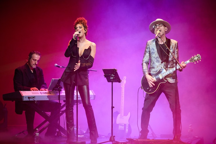 Drei Musiker stehen auf der Bühne: links ein Keyboarder, in der Mitte eine Sängerin mit Mikrofon und rechts ein Gitarrist mit Hut; sie sind von einer lila-rosa Bühnenbeleuchtung umgeben.