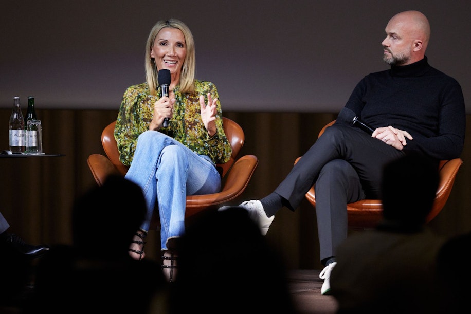 A woman holding a microphone speaks while sitting on stage next to a man in a black sweater; both are seated in brown chairs with bottles of water on a side table beside them. Audience silhouettes are visible in the foreground.