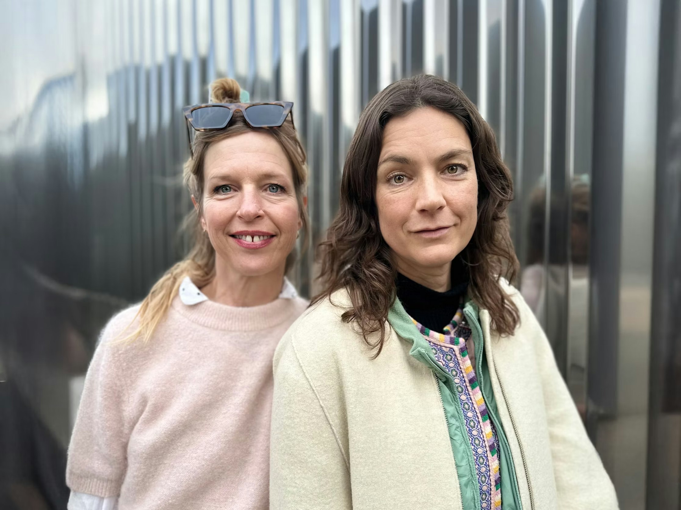 Zwei Frauen stehen dicht beieinander draußen und lächeln sanft in die Kamera. Die eine trägt eine Sonnenbrille und einen hellrosa Pullover; die andere hat welliges braunes Haar und eine cremefarbene Jacke mit einem bunten Oberteil. Im Hintergrund ist eine Metallwand zu sehen.