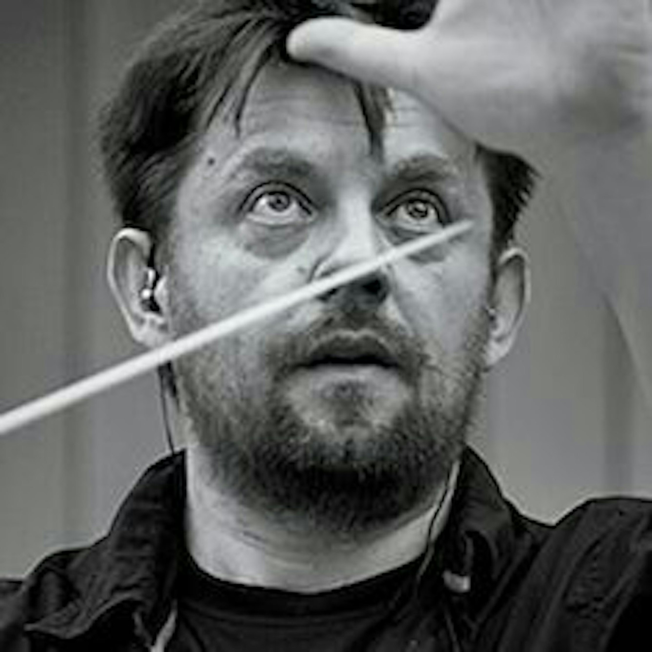 Ein Mann mit Bart und Kopfhörern blickt nach oben, während er die Hand mit einem Taktstock in der Hand hebt. Das Bild ist schwarz-weiß.