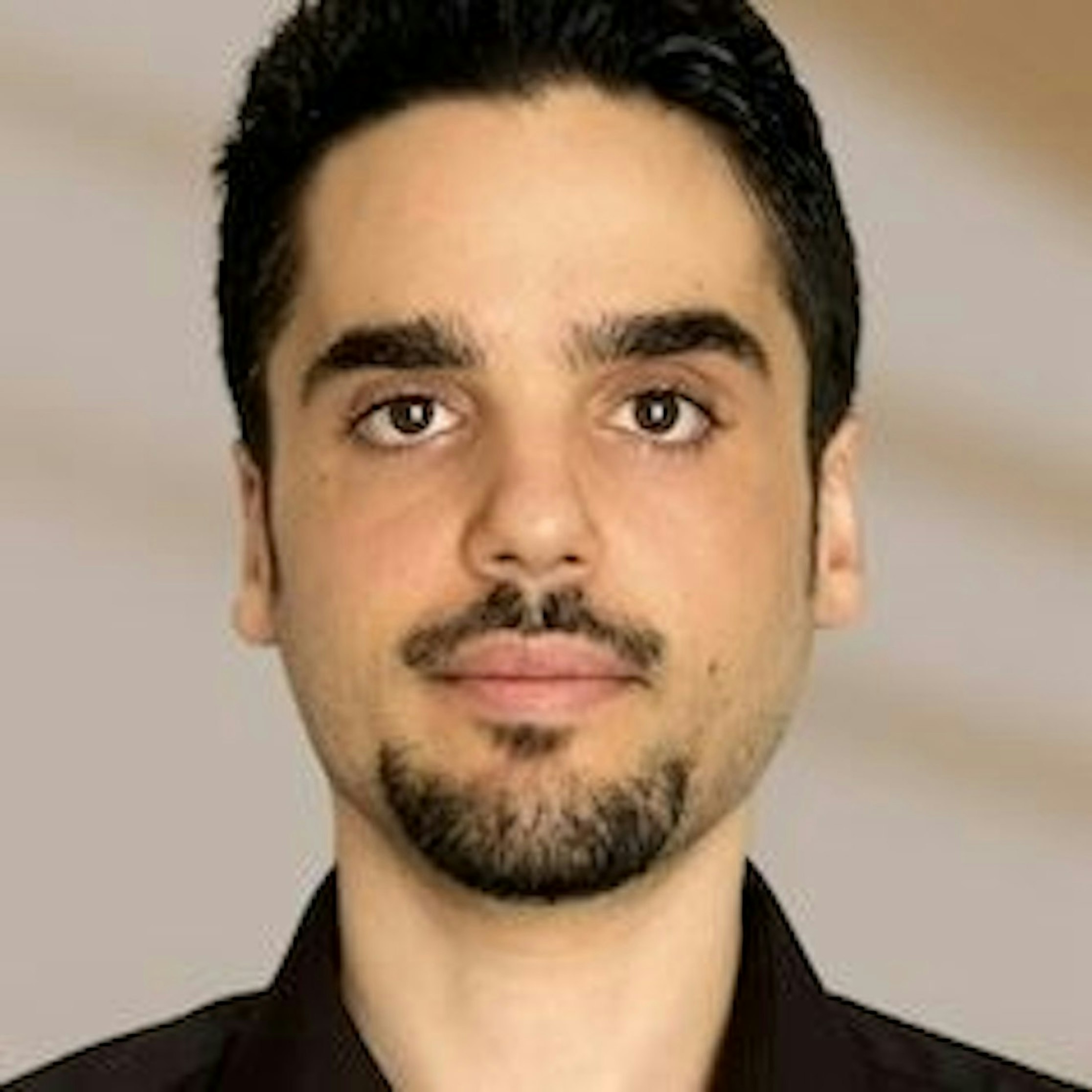 Ein Mann mit kurzen dunklen Haaren, gestutztem Voll- und Schnurrbart blickt in die Kamera. Er trägt ein dunkles Hemd und steht vor einem unscharfen, hellbraunen Hintergrund.