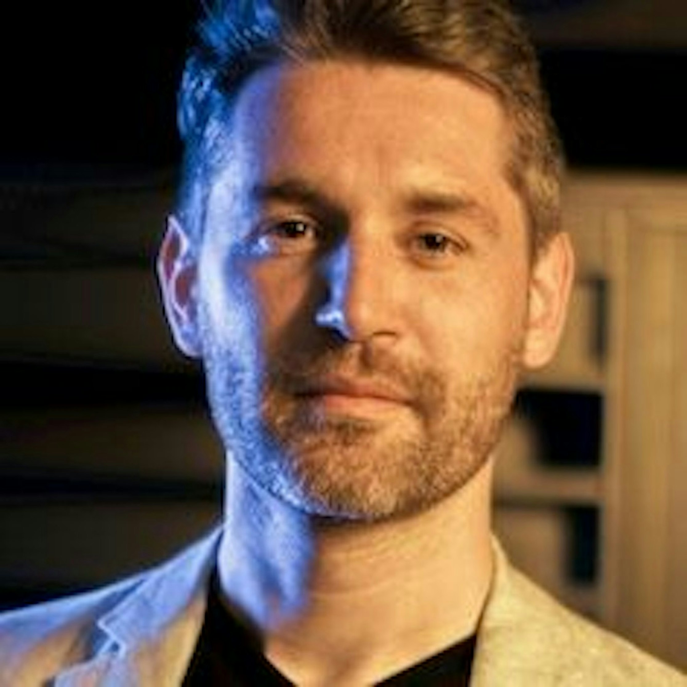 Ein Mann mit kurzen braunen Haaren und gestutztem Bart trägt eine beige Jacke über einem dunklen Hemd und blickt mit einem leichten Lächeln in die Kamera. Die Beleuchtung ist warm und blau.