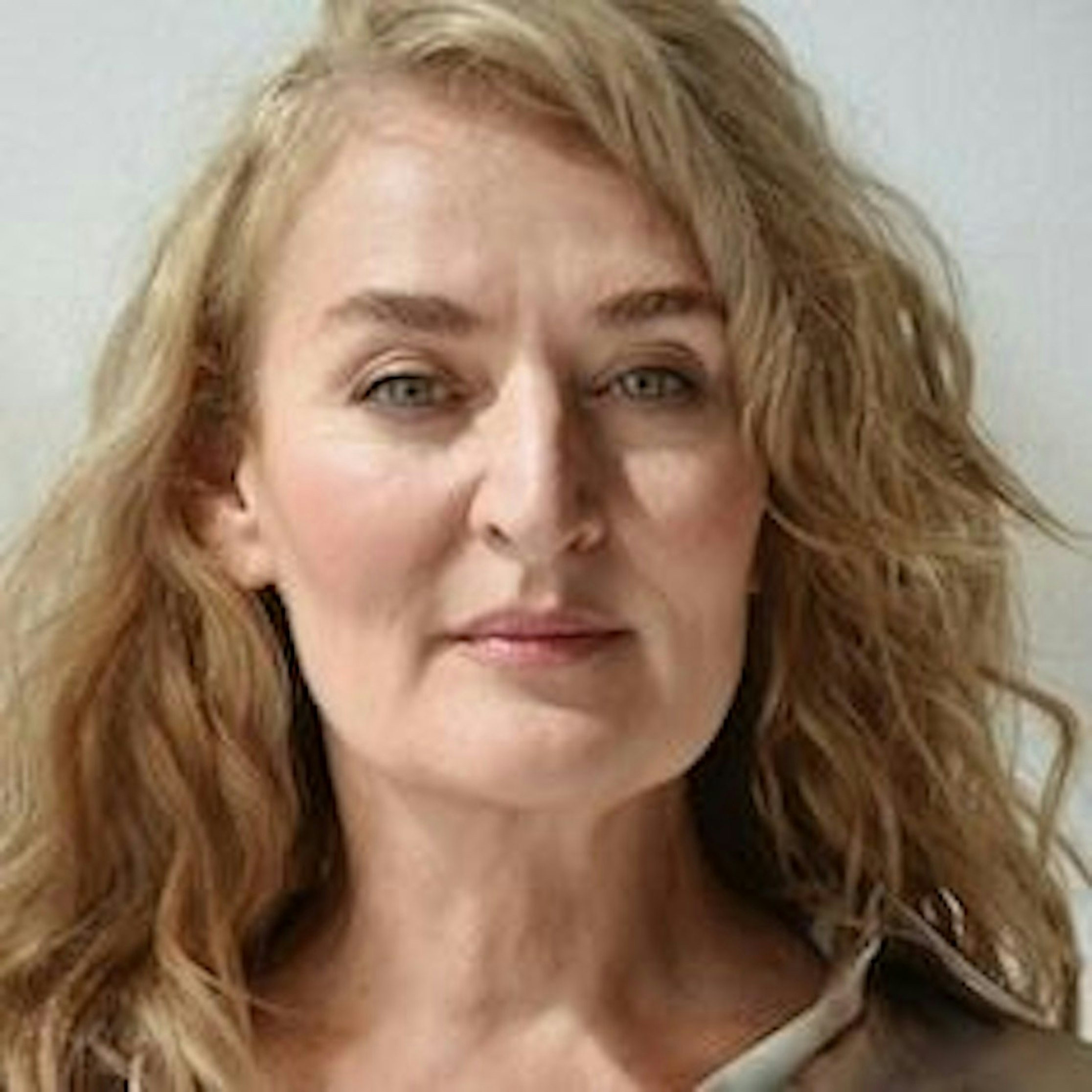 Eine Frau mit welligem blondem Haar und heller Haut blickt mit neutralem Gesichtsausdruck direkt in die Kamera und steht vor einem schlichten hellen Hintergrund.
