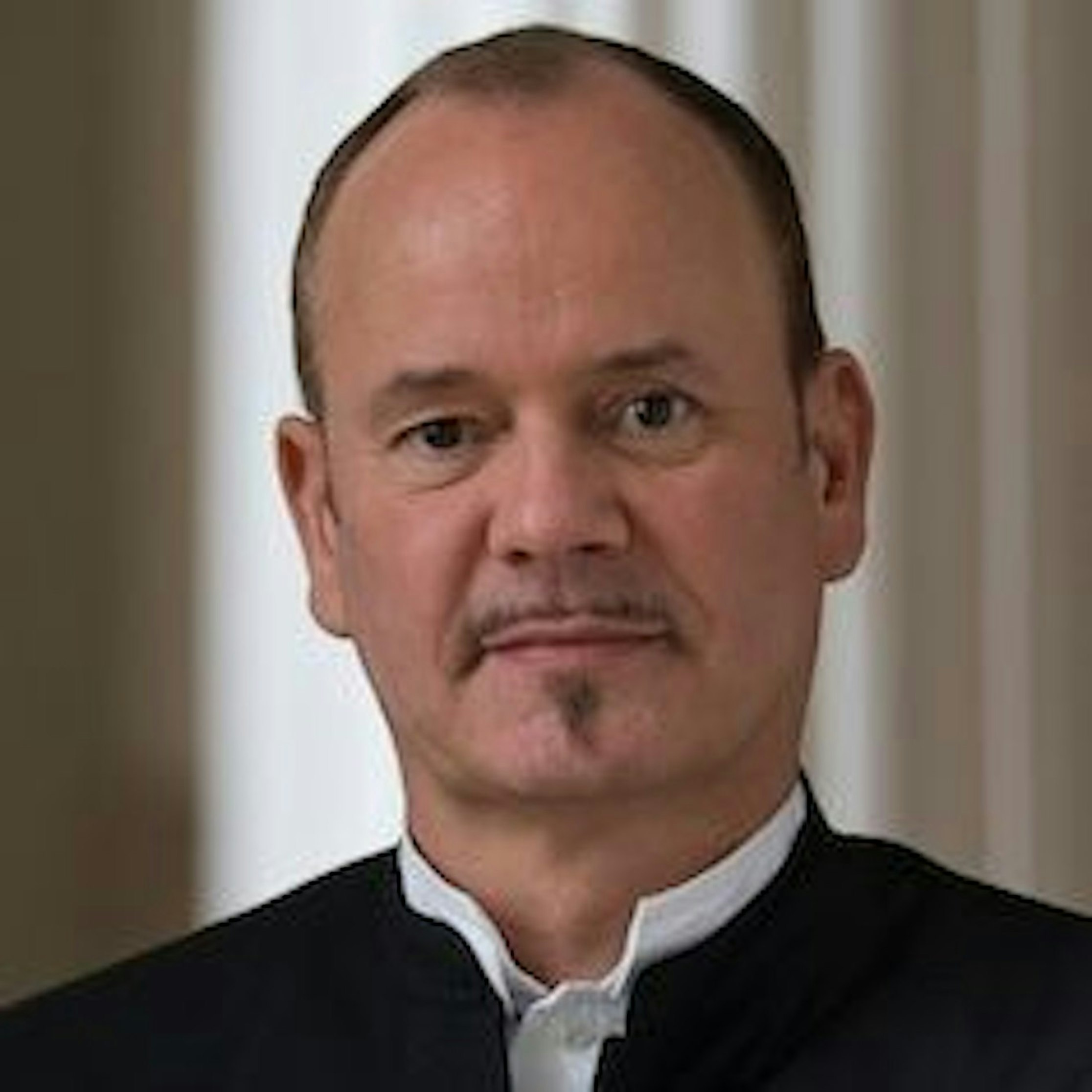 Ein Mann mittleren Alters mit zurückweichendem Haaransatz, heller Haut und gestutztem Schnurrbart und Spitzbart, der eine dunkle Jacke über einem weißen Hemd trägt, steht vor verschwommenen weißen Säulen.