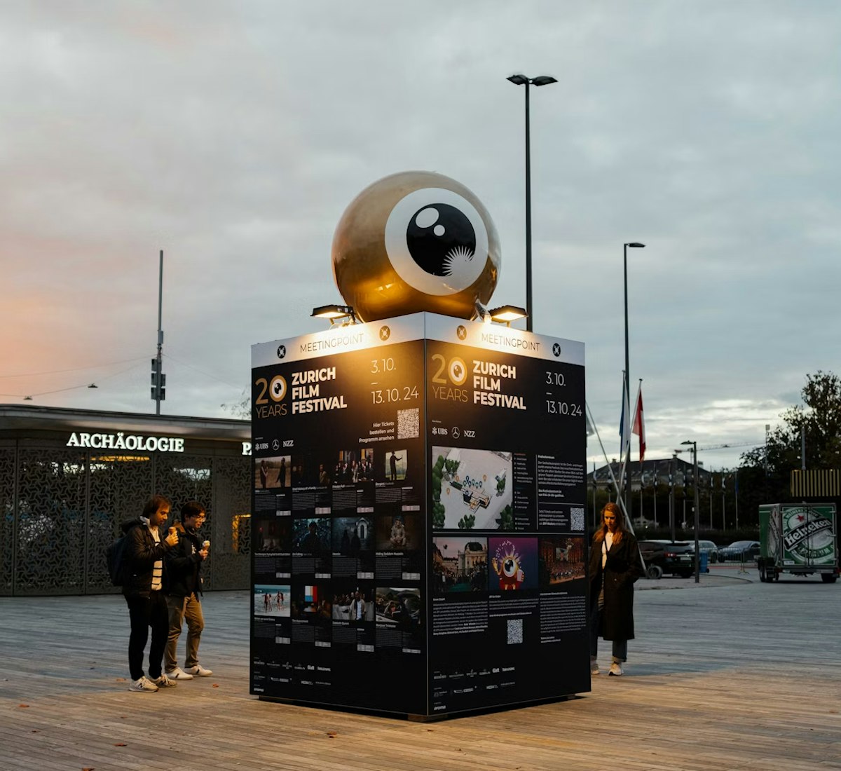 festival_zentrum Ein paar Leute stehen bei Sonnenuntergang auf einem Aussenplatz neben einem grossen, beleuchteten Kubus mit Filmplakaten und einer riesigen Augapfel-Skulptur darauf, die für das Zurich Film Festival wirbt.