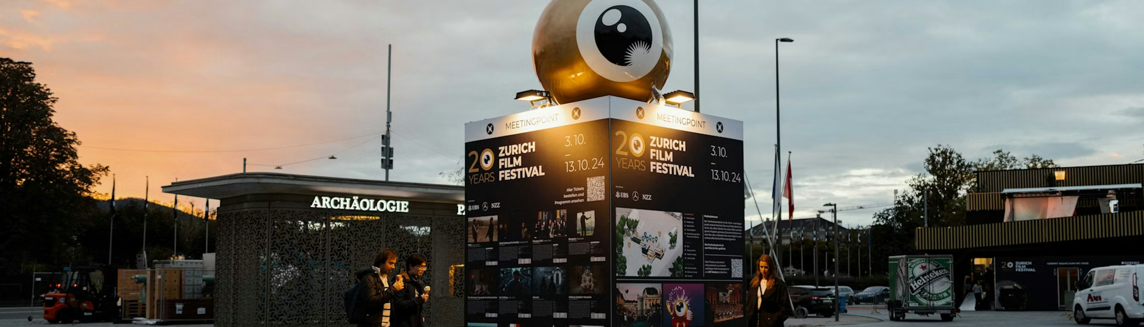festival_zentrum Ein paar Leute stehen bei Sonnenuntergang auf einem Aussenplatz neben einem grossen, beleuchteten Kubus mit Filmplakaten und einer riesigen Augapfel-Skulptur darauf, die für das Zurich Film Festival wirbt.