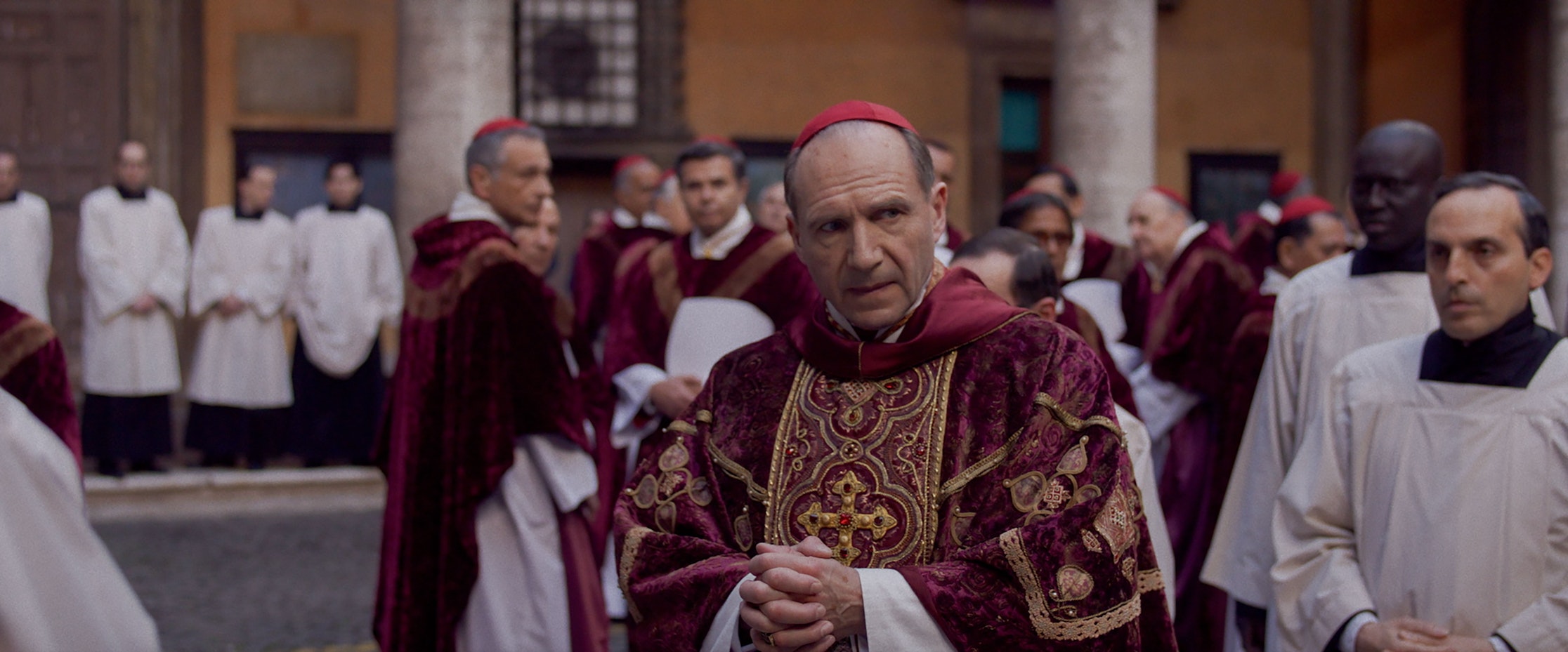 Eine Gruppe Geistlicher in reich verzierten kastanienbraunen und goldenen Gewändern steht vor einem Gebäude. Im Vordergrund steht ein ernst dreinblickender Kardinal. In der Gruppe sind auch Priester in weißen Gewändern anwesend.