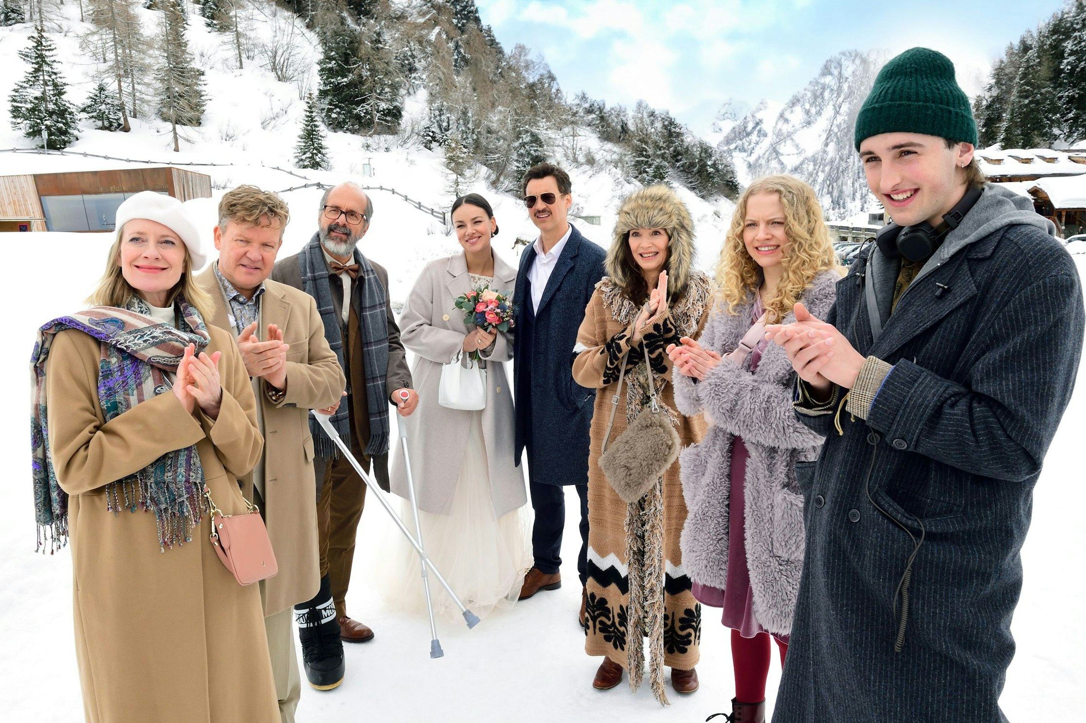 Eine Gruppe von Menschen in Winterkleidung steht im Freien in einer verschneiten Berglandschaft und klatscht; in der Mitte stehen Braut und Bräutigam, umgeben von lächelnden Freunden und Verwandten.