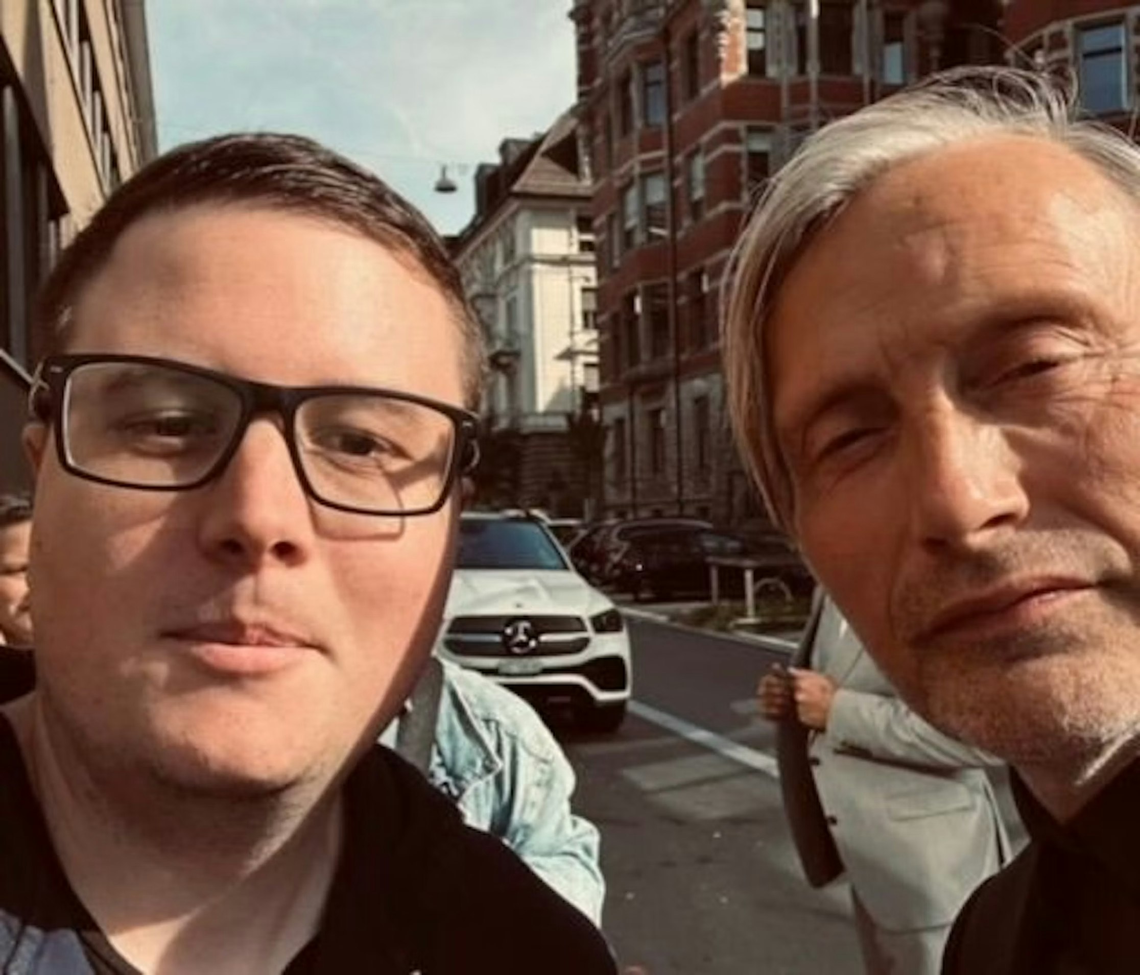 Zwei Männer machen gemeinsam ein Selfie auf einer Stadtstraße. Im Hintergrund sind Gebäude und ein weißer Mercedes-Benz zu sehen. Einer trägt eine Brille, der andere hat graue Haare und blinzelt in die Kamera.