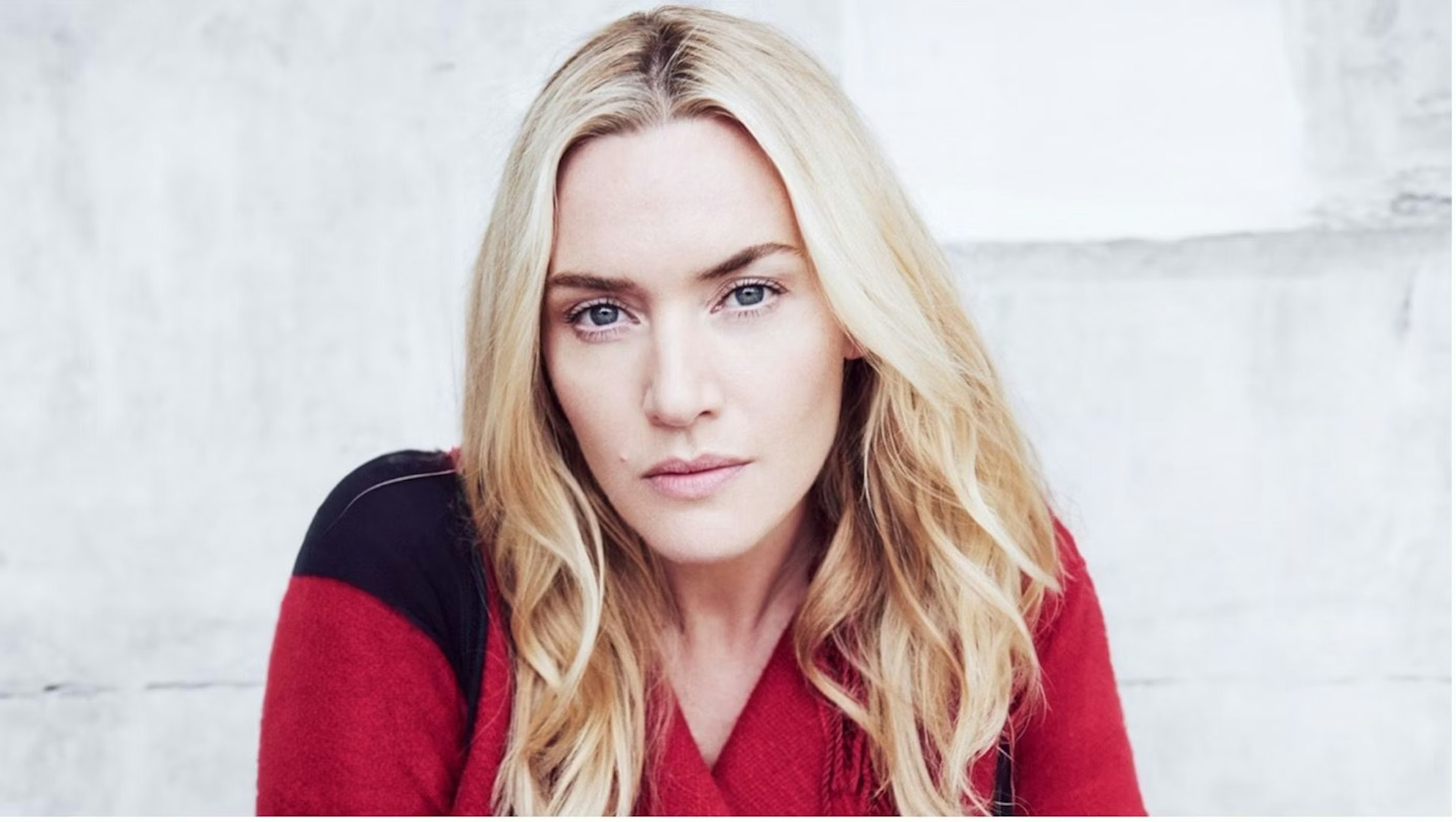 Eine Frau mit langen blonden Haaren und einem rot-schwarzen Pullover blickt mit neutralem Gesichtsausdruck vor einem hellgrauen Hintergrund direkt in die Kamera.