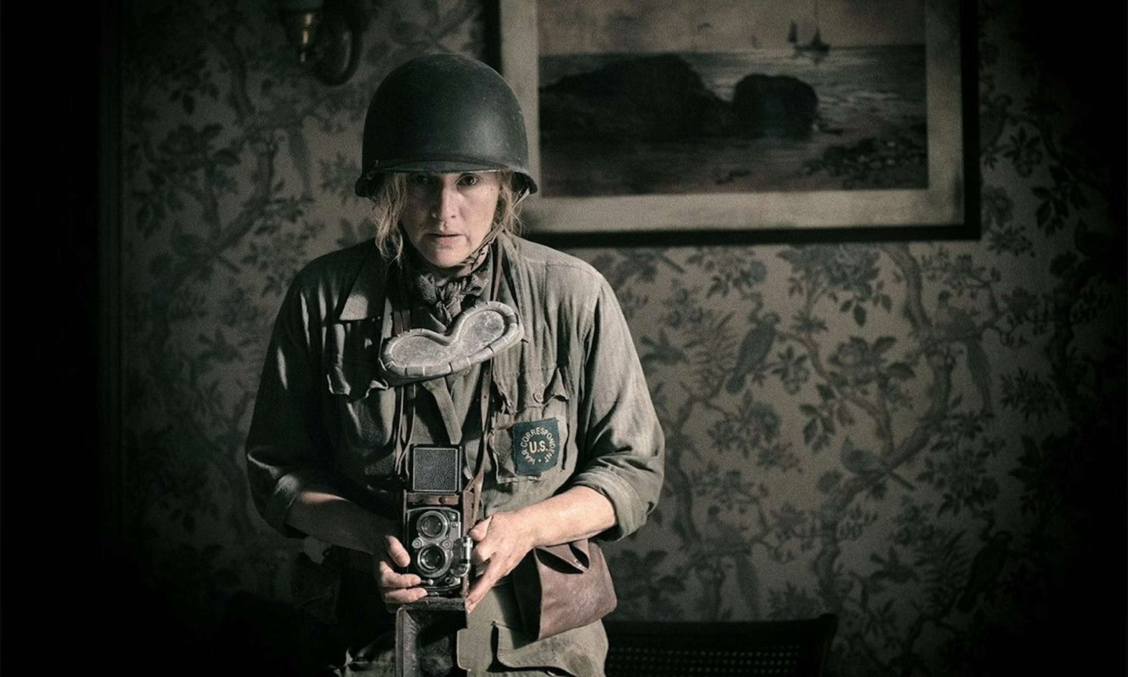Eine Person in Militärhelm und Uniform hält eine altmodische Kamera in der Hand. Sie steht in einem Raum vor einer gemusterten Wand, hinter ihr hängt ein Gemälde mit einer Meereslandschaft. Die Beleuchtung ist gedämpft und stimmungsvoll.