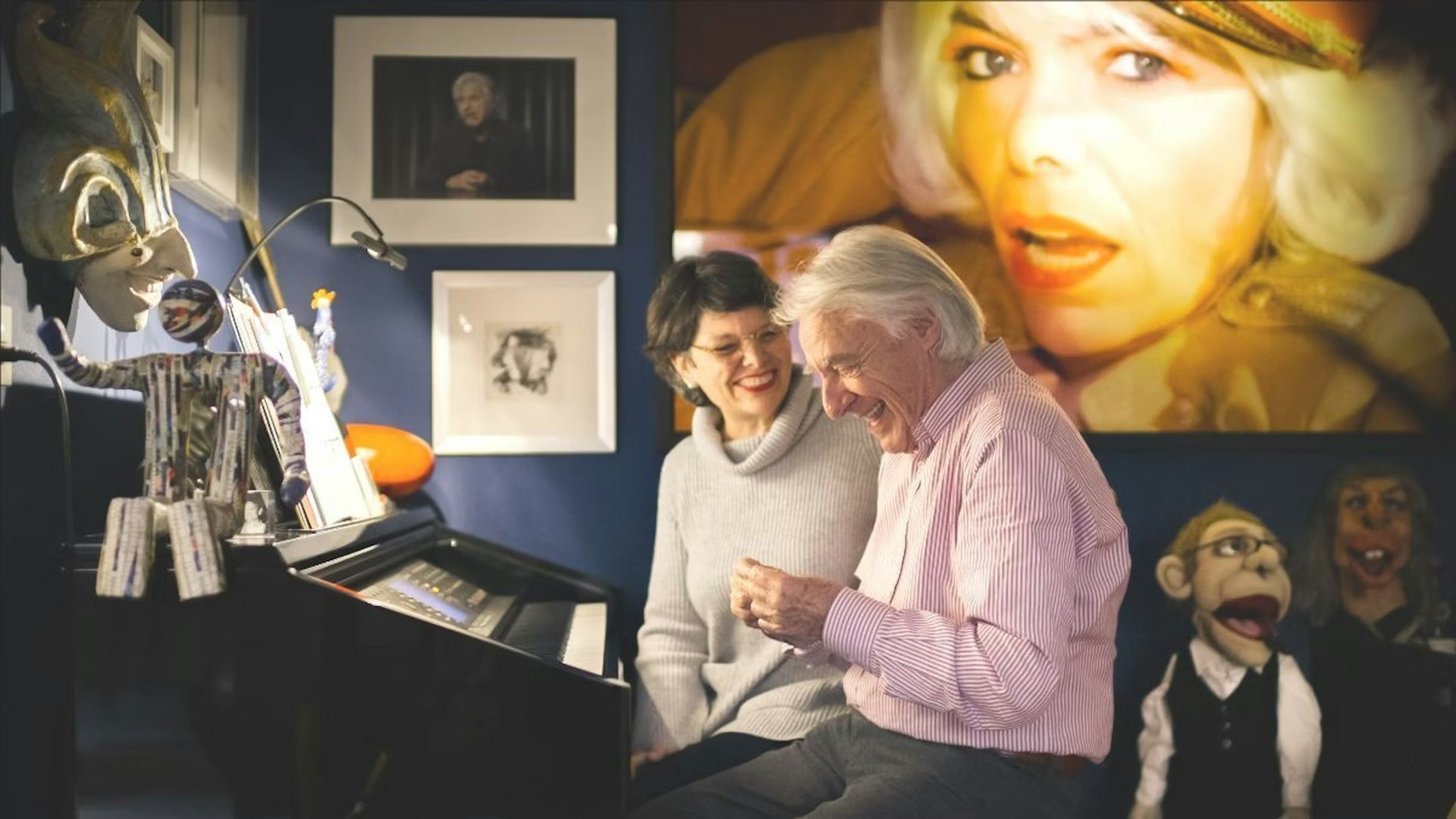 Ein älterer Mann und eine Frau sitzen lächelnd an einem Klavier in einem farbenfrohen, kunstvollen Raum. Dahinter befinden sich gerahmte Fotos, ein großes Frauenporträt sowie mehrere Puppen und Figuren, die eine lebendige, kreative Atmosphäre schaffen.