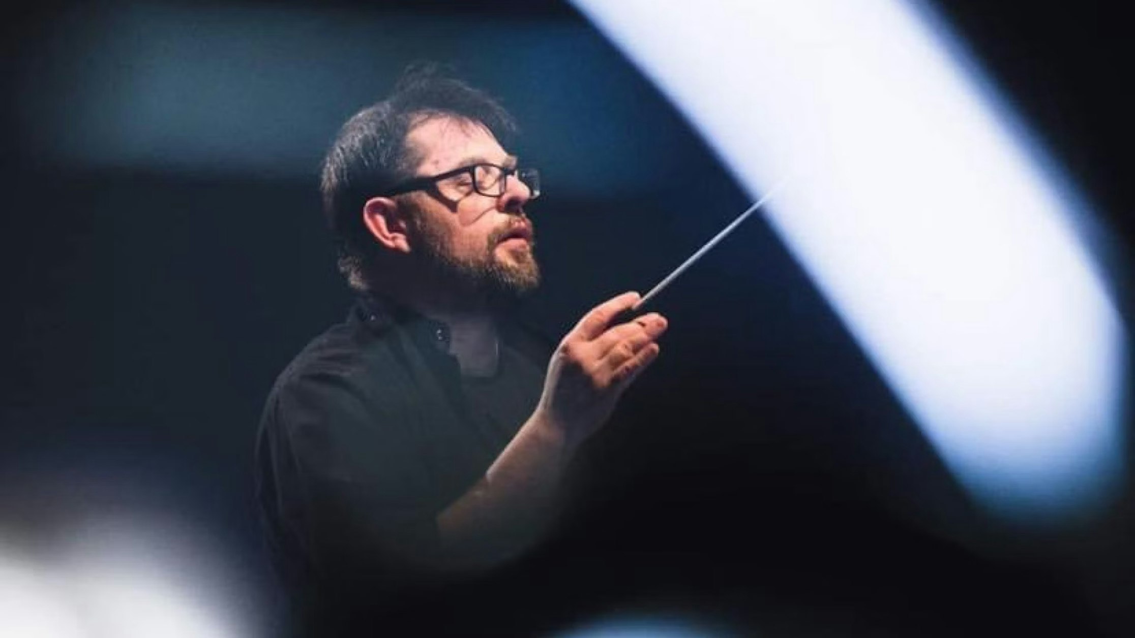 Ein bärtiger Mann mit Brille hält einen Dirigentenstab in der Hand und gestikuliert, als würde er ein Orchester leiten. Weiche, unscharfe Lichtstreifen rahmen den dunklen, dramatischen Hintergrund ein.