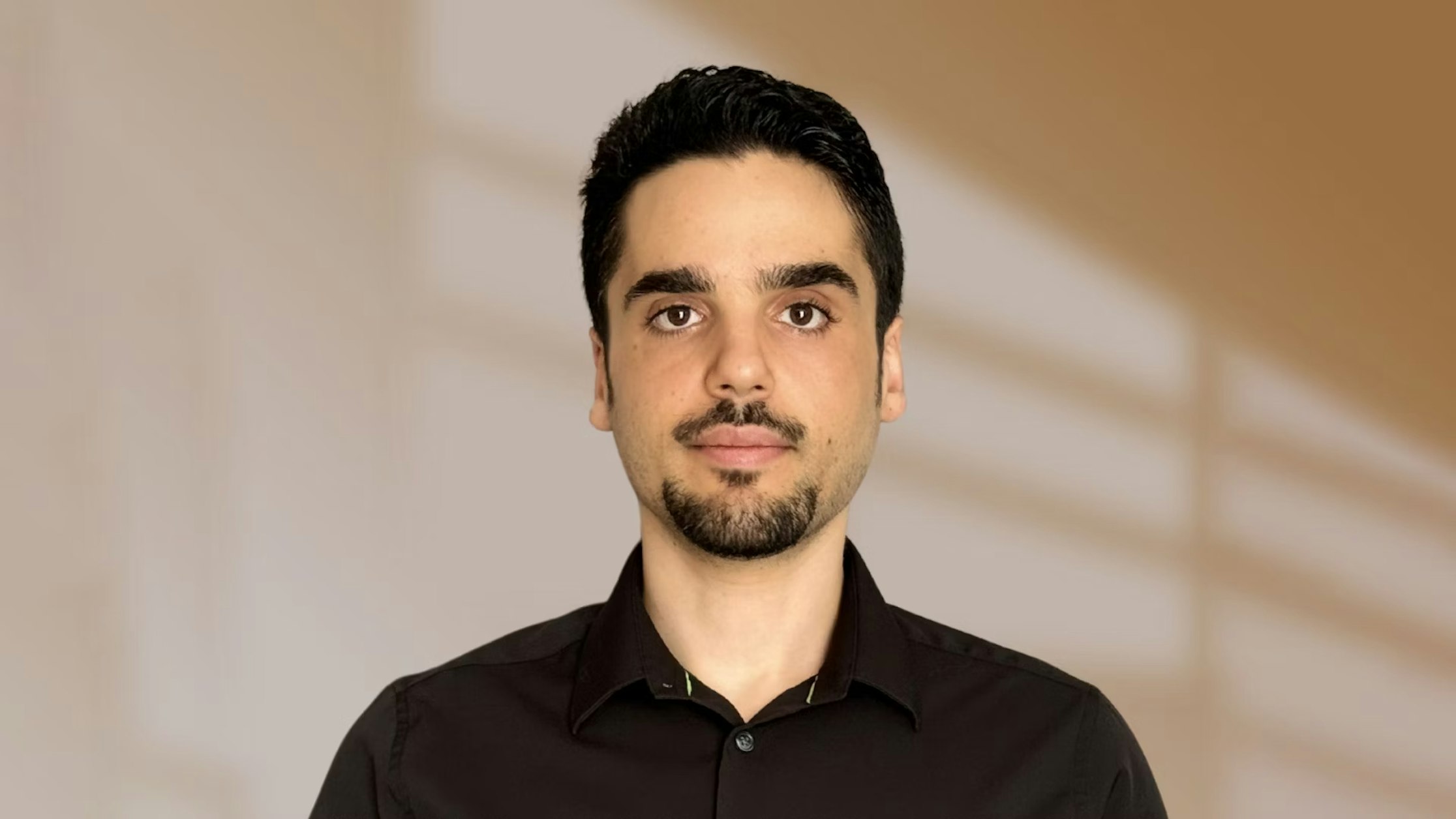 Ein Mann mit kurzen dunklen Haaren und gestutztem Bart, der ein schwarzes Hemd mit Knöpfen trägt, steht vor einem unscharfen beigen Hintergrund mit weichen Linien.