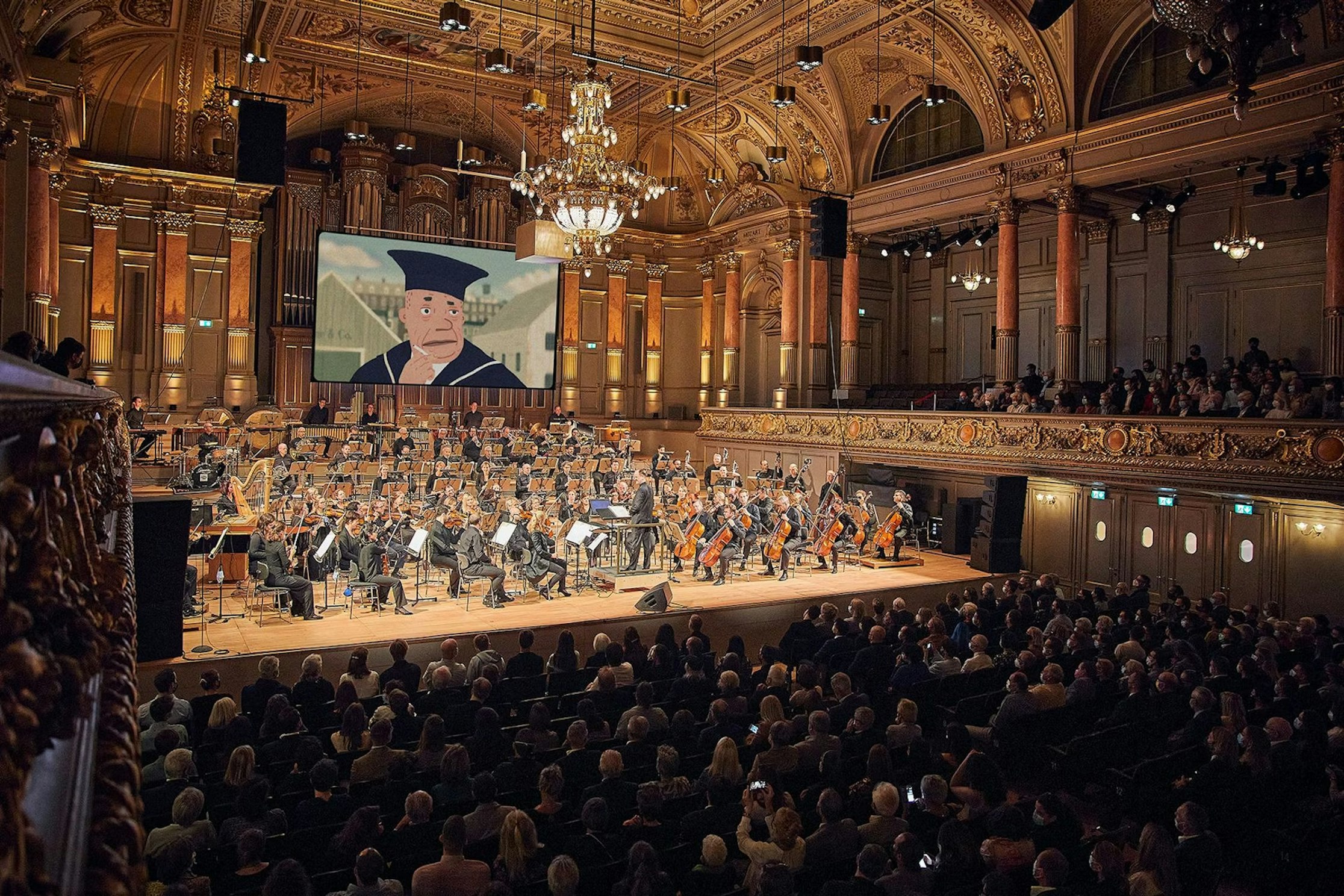 Auf der Bühne eines reich verzierten Konzertsaals tritt ein komplettes Orchester auf. Darüber ist auf einer großen Leinwand eine Zeichentrickfigur in akademischer Robe zu sehen, während ein sitzendes Publikum zusieht.