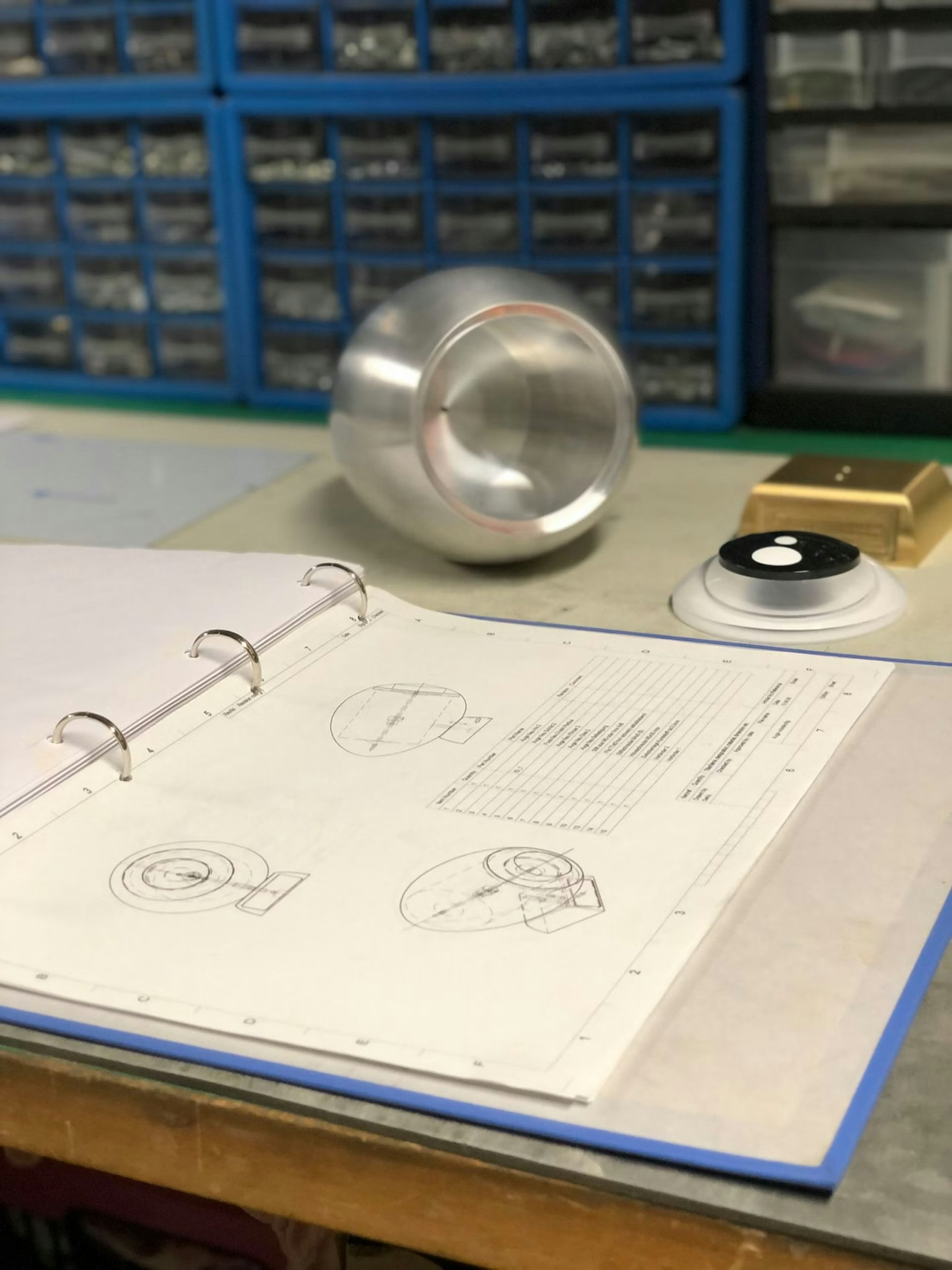 Auf einem Schreibtisch liegt ein geöffneter Ordner mit technischen Zeichnungen neben einem zylindrischen Metallobjekt, einem runden Knopfgerät und kleinen Schubladen mit geordneten Teilen im Hintergrund.