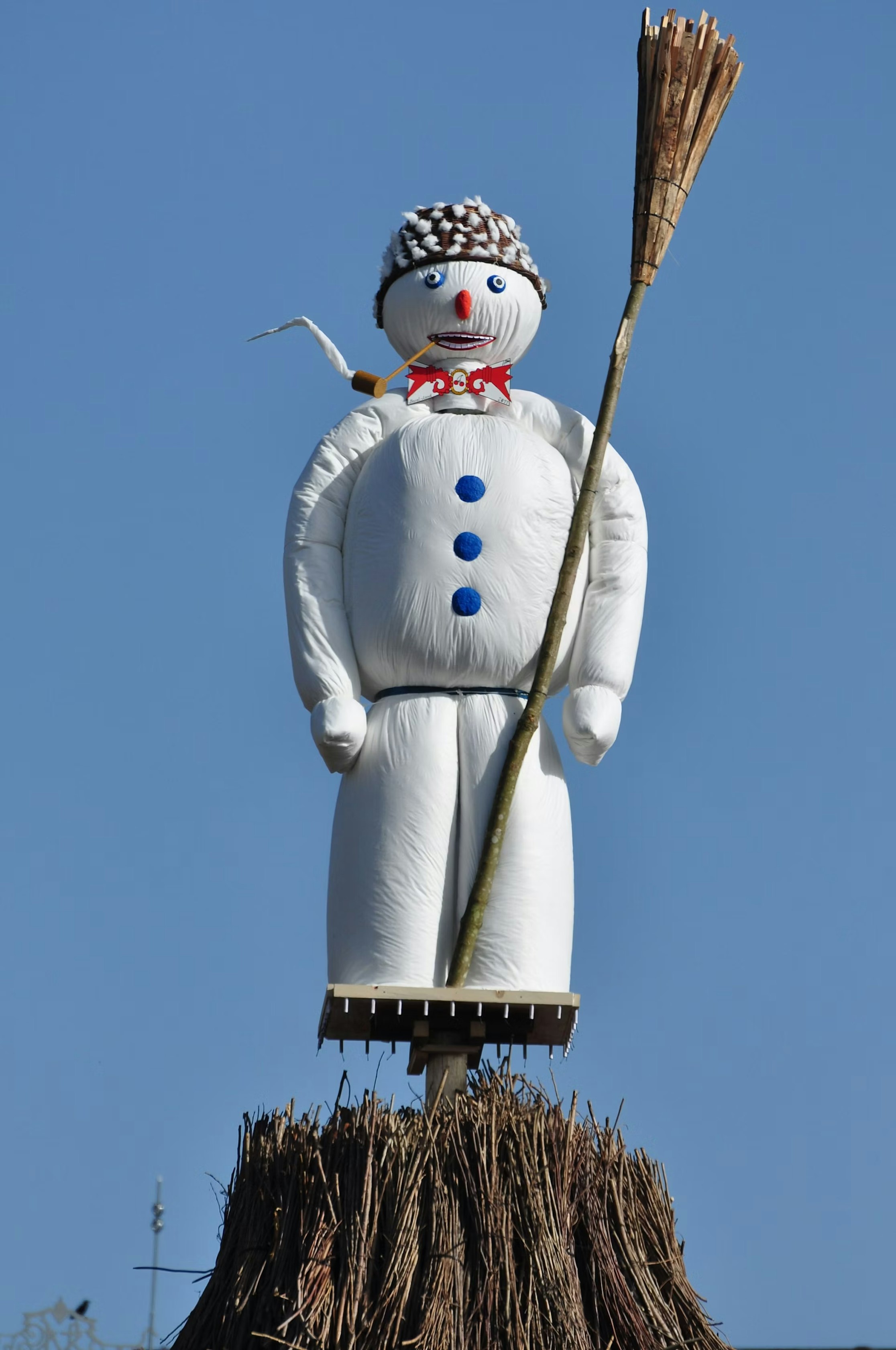 Eine große Schneemannfigur mit blauen Knöpfen, einer roten Fliege und einem Hut steht auf einem Bündel Stöcke und hält einen Besen vor einem klaren blauen Himmel.