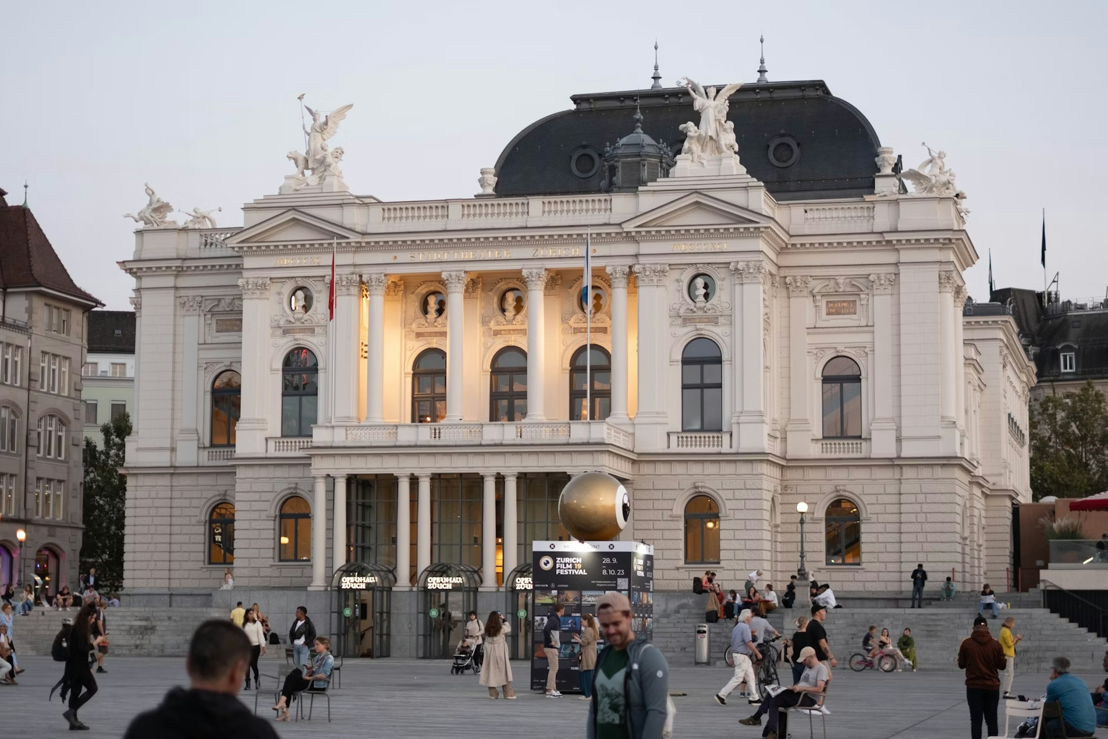Bei Tageslicht versammeln sich Menschen auf einem belebten Stadtplatz vor dem kunstvollen Opernhaus Zürich mit seinen imposanten Säulen, Statuen und dekorativen Details an der Fassade.