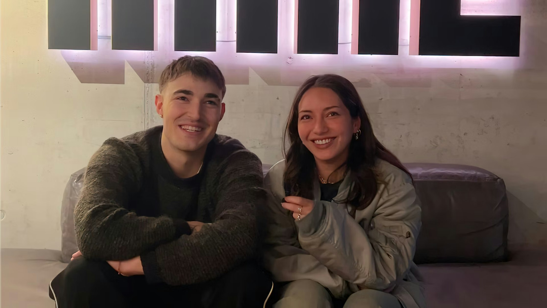 Zwei Personen sitzen auf einem Sofa und lächeln in die Kamera. Die linke Person trägt einen dunklen Pullover, die rechte eine graue Jacke. Hinter ihnen leuchtet ein Neonschild.