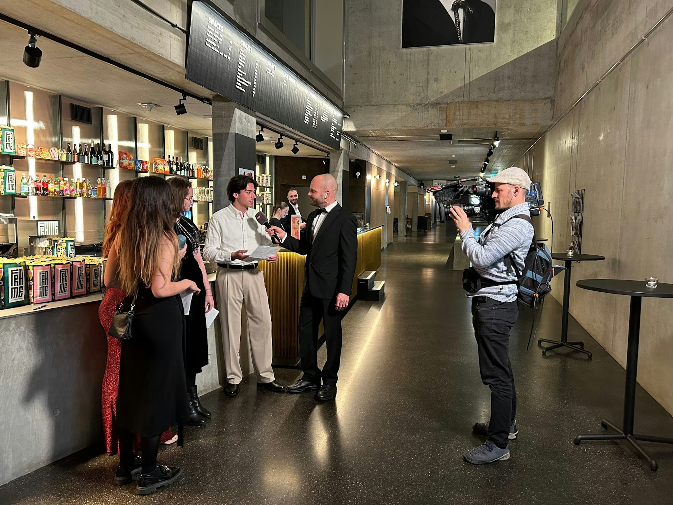 Ein Kameramann filmt eine kleine Gruppe von Menschen, die sich in einer modernen Bar oder Lobby im Industriestil mit Betonwänden und beleuchtetem Tresen unterhalten und Papiere halten. Ein Mann im Anzug scheint einen anderen Mann zu interviewen.